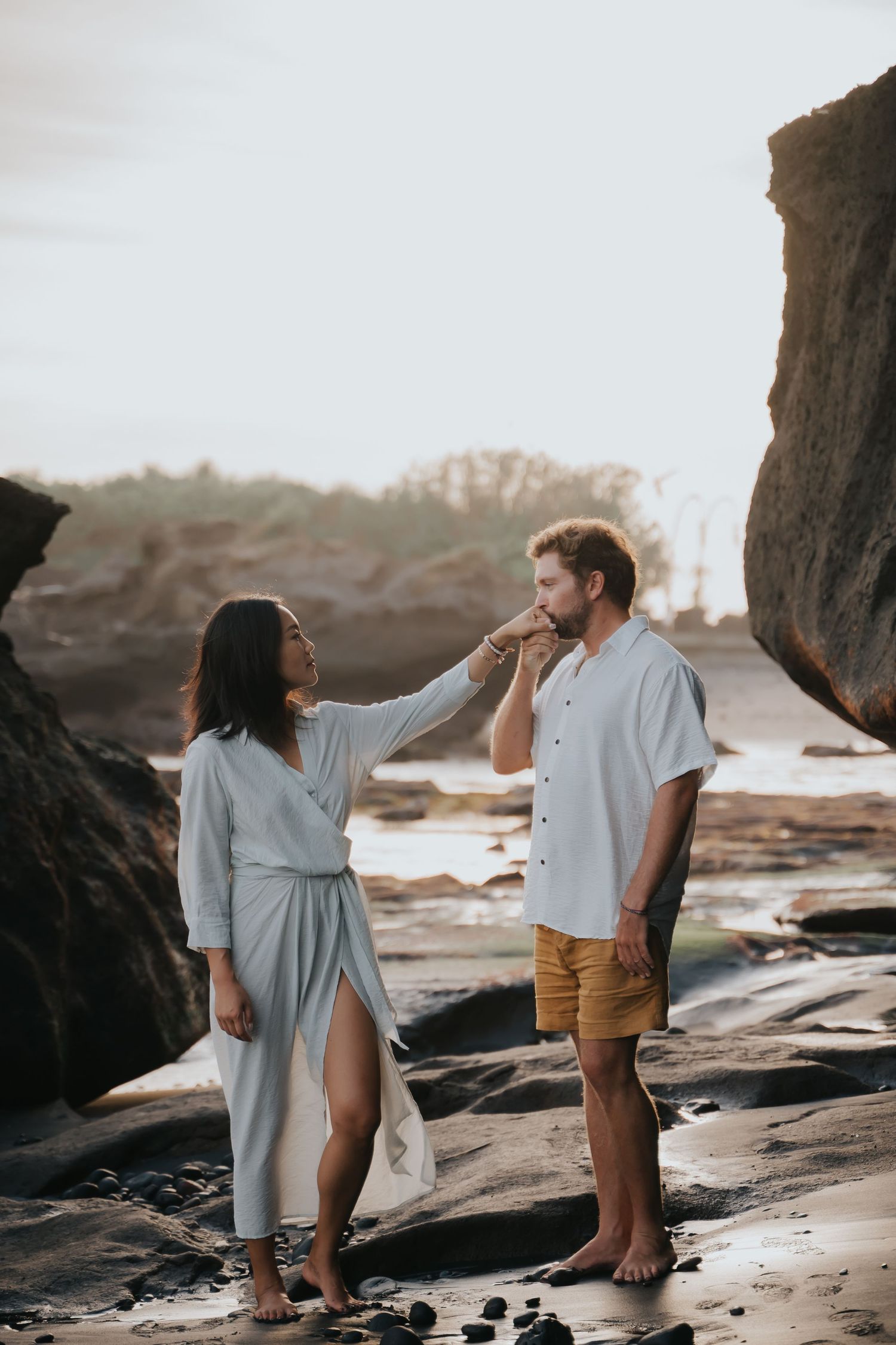 Couple walking barefoot on rocky beach during sunset while holding hands and exploring coastal scenery.