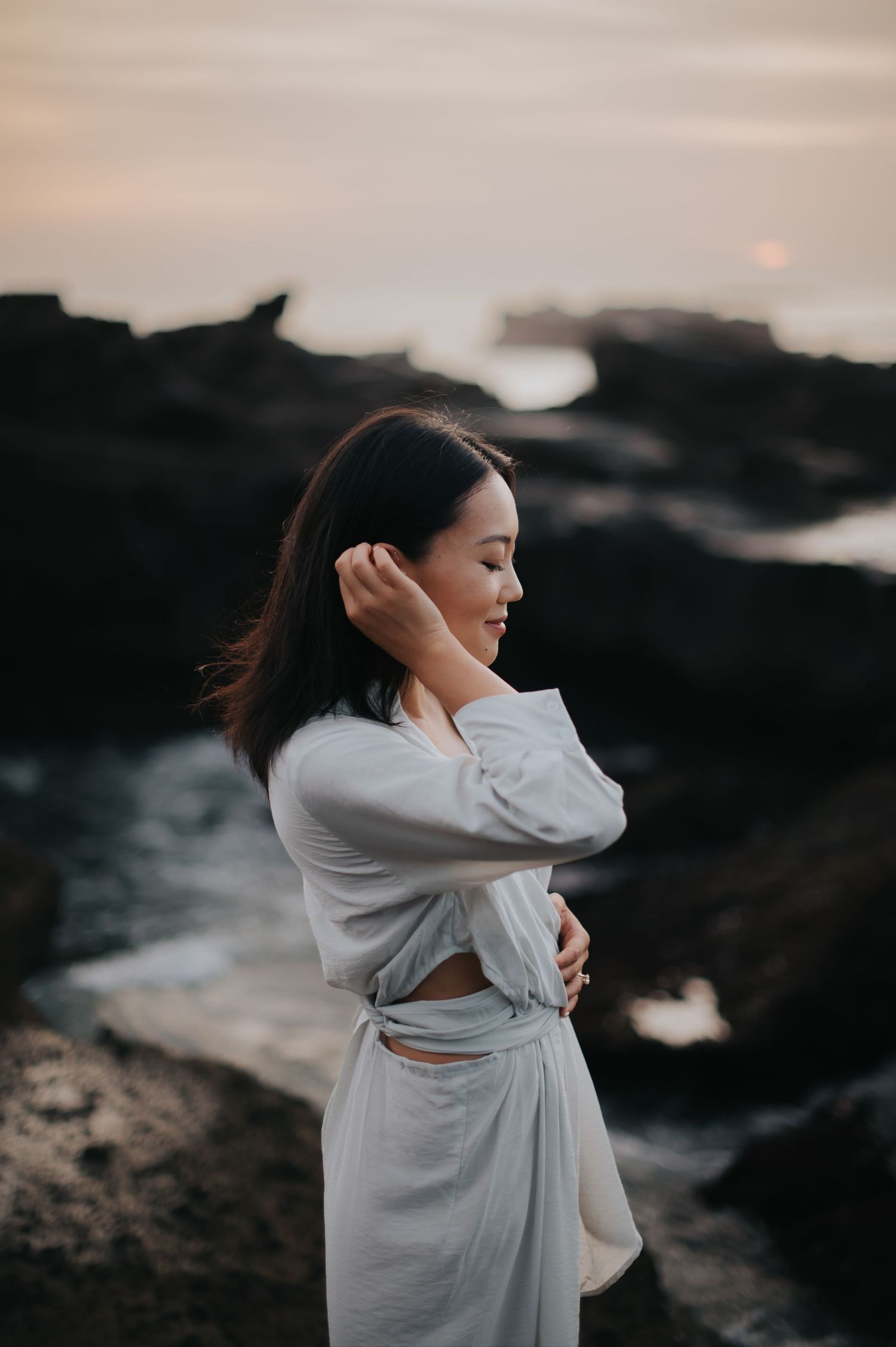 A person in a flowing white dress poses gracefully against a dramatic rocky coastal landscape at sunset.