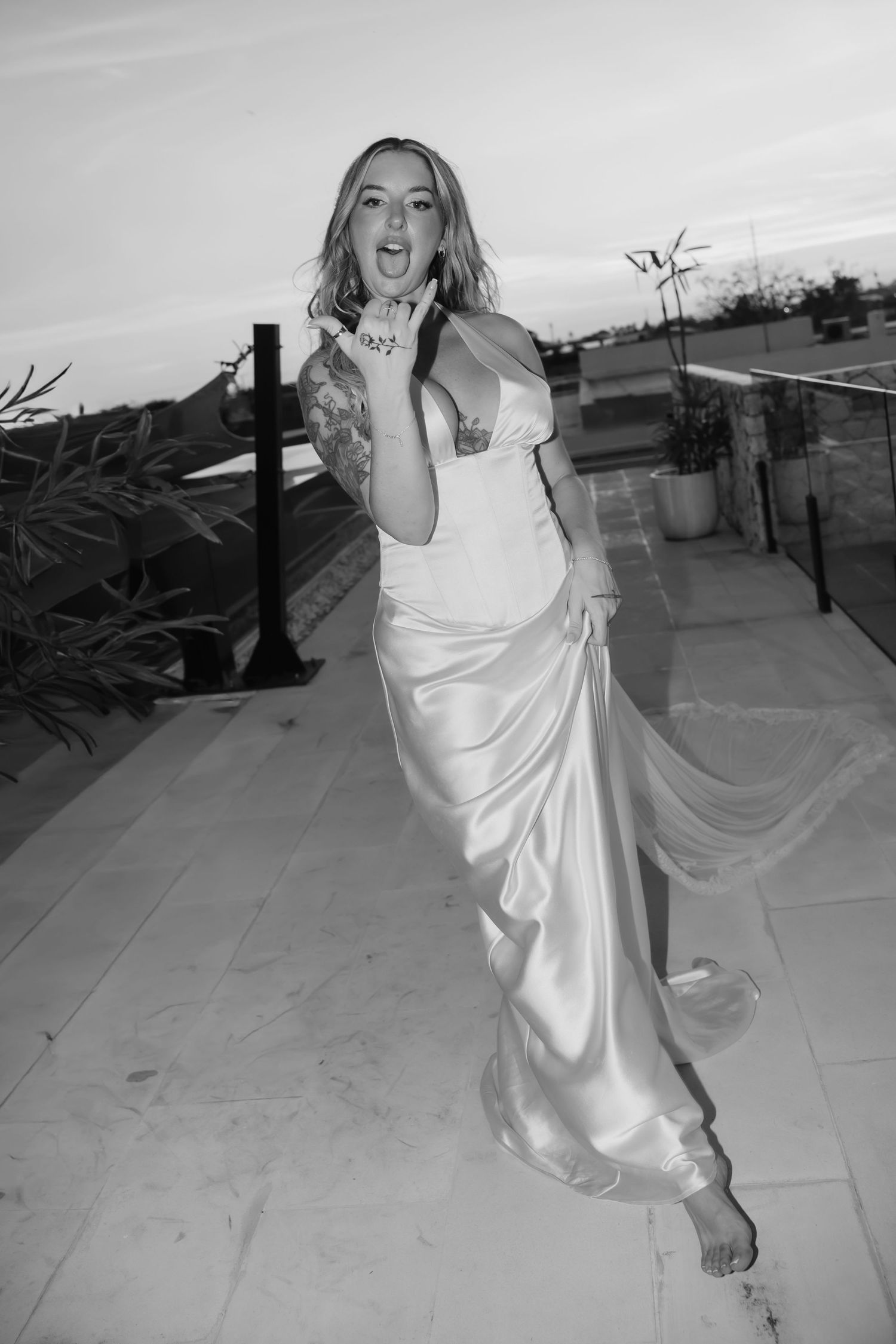 Bride in a satin wedding gown poses barefoot on a rooftop terrace at dusk in a joyful black and white photo.