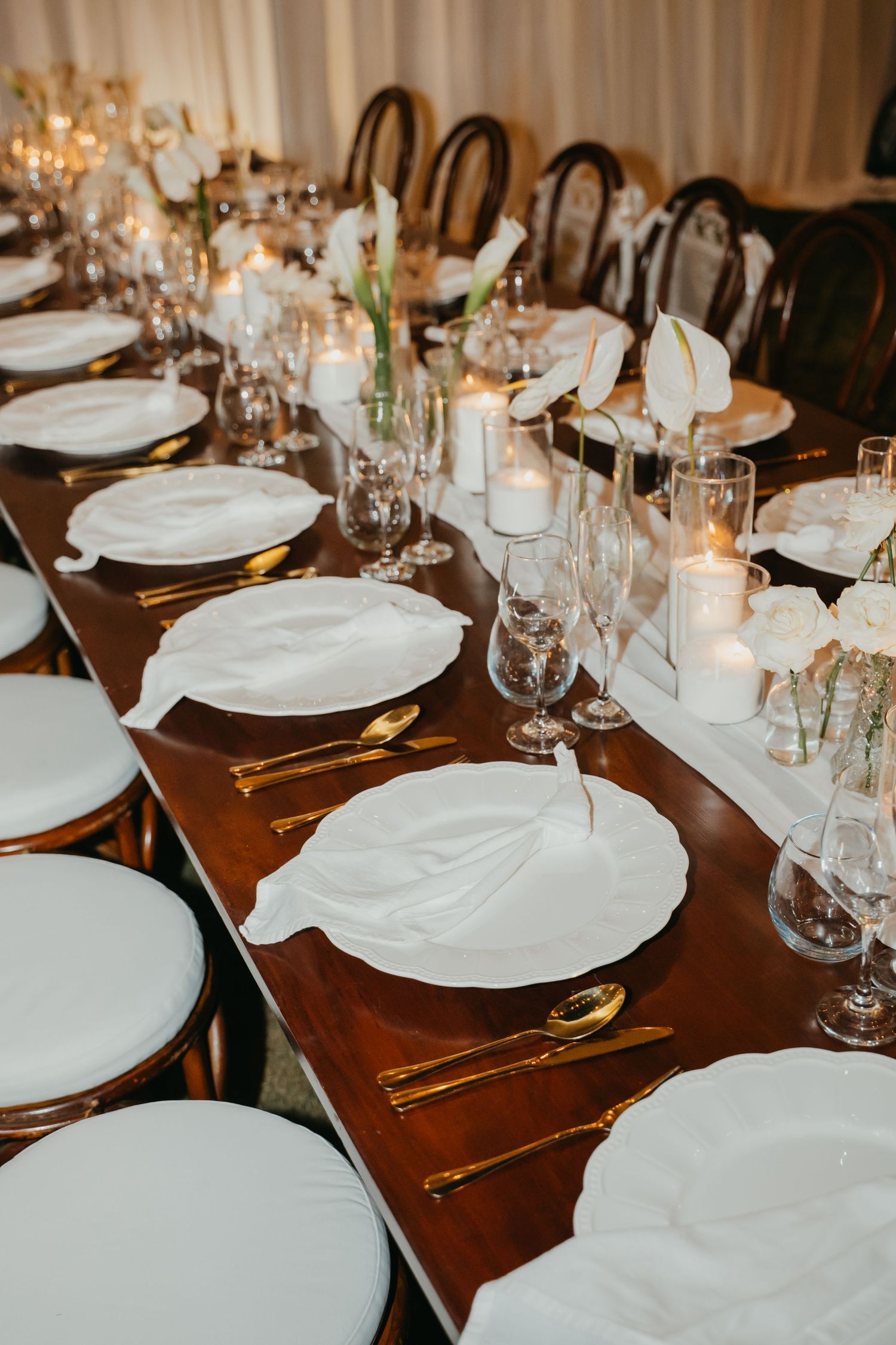 Elegant wedding reception table with white plates, gold cutlery, crystal glassware, white floral centerpieces, and candles on dark wood.