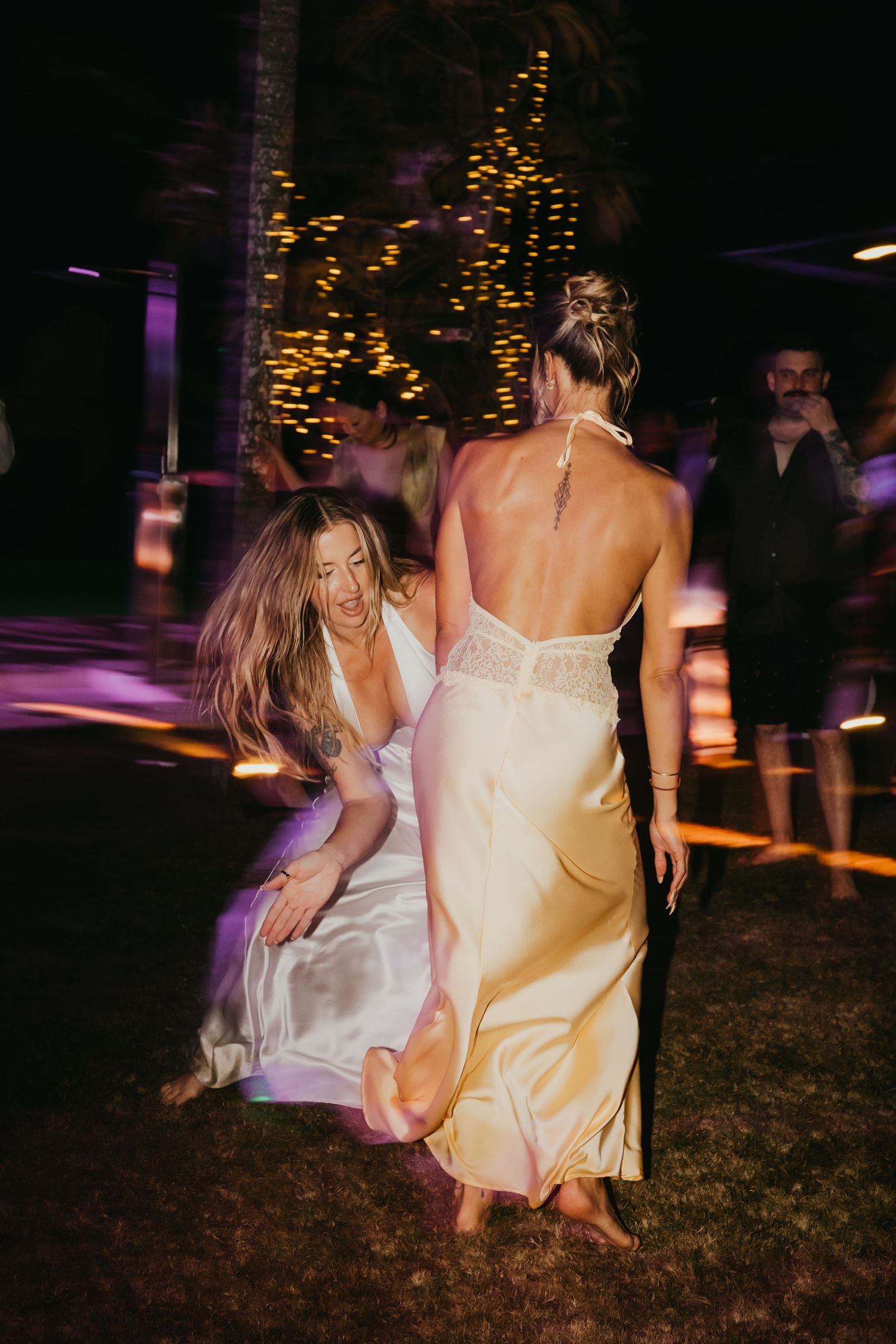 Two women in elegant satin gowns dance energetically at an outdoor nighttime wedding celebration.