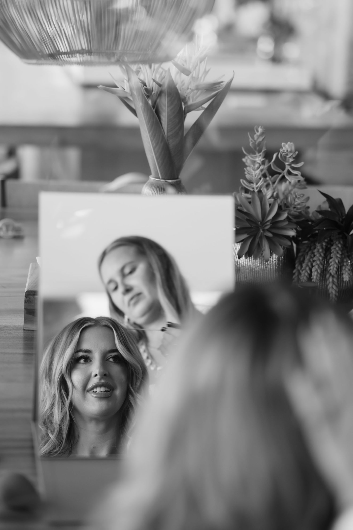 Black and white photo of a blonde bride admiring her reflection in a mirror surrounded by floral arrangements.