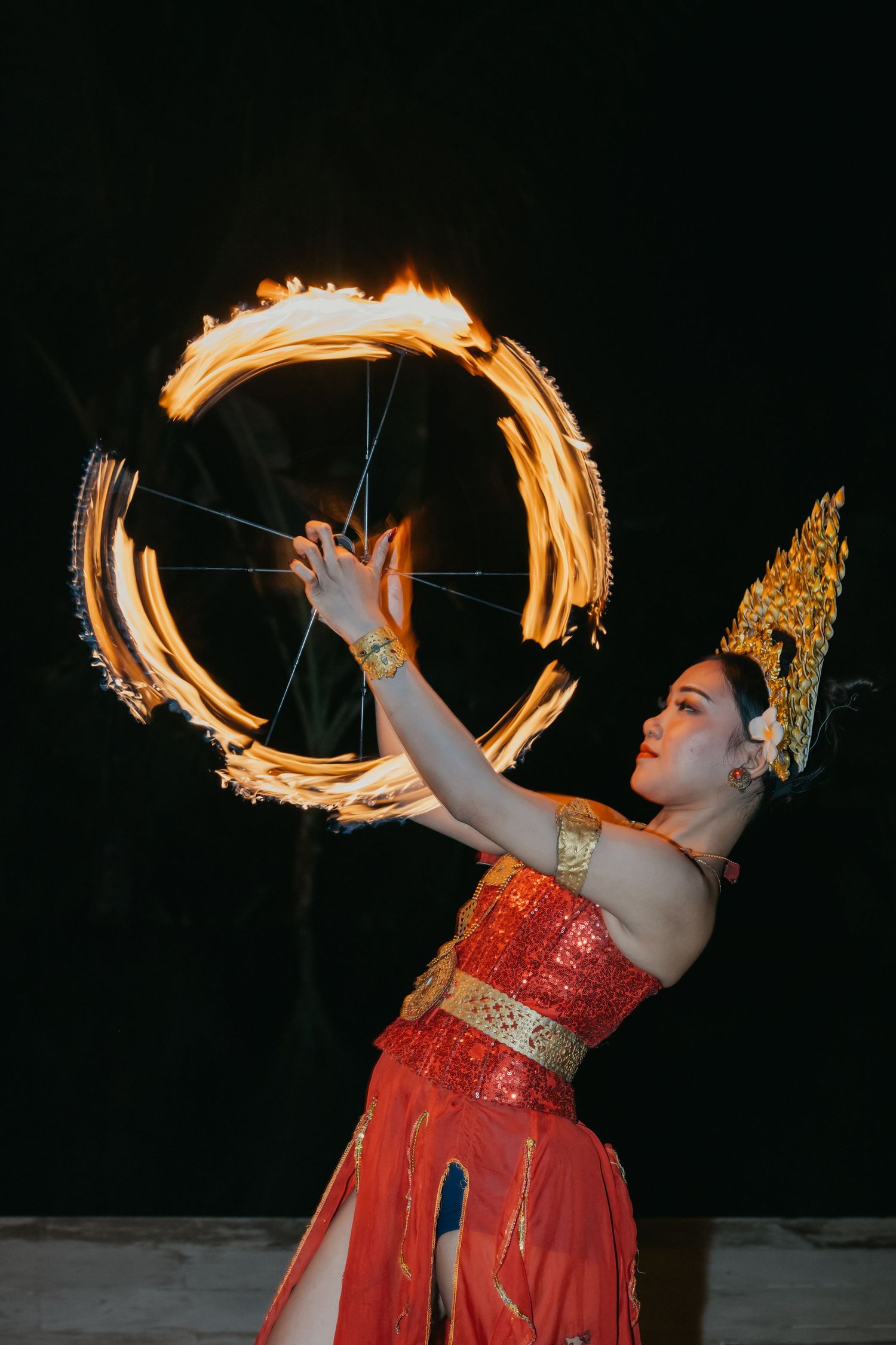 Traditional dancer in red costume performs mesmerizing fire spinning creating circular flame patterns against dark night sky.
