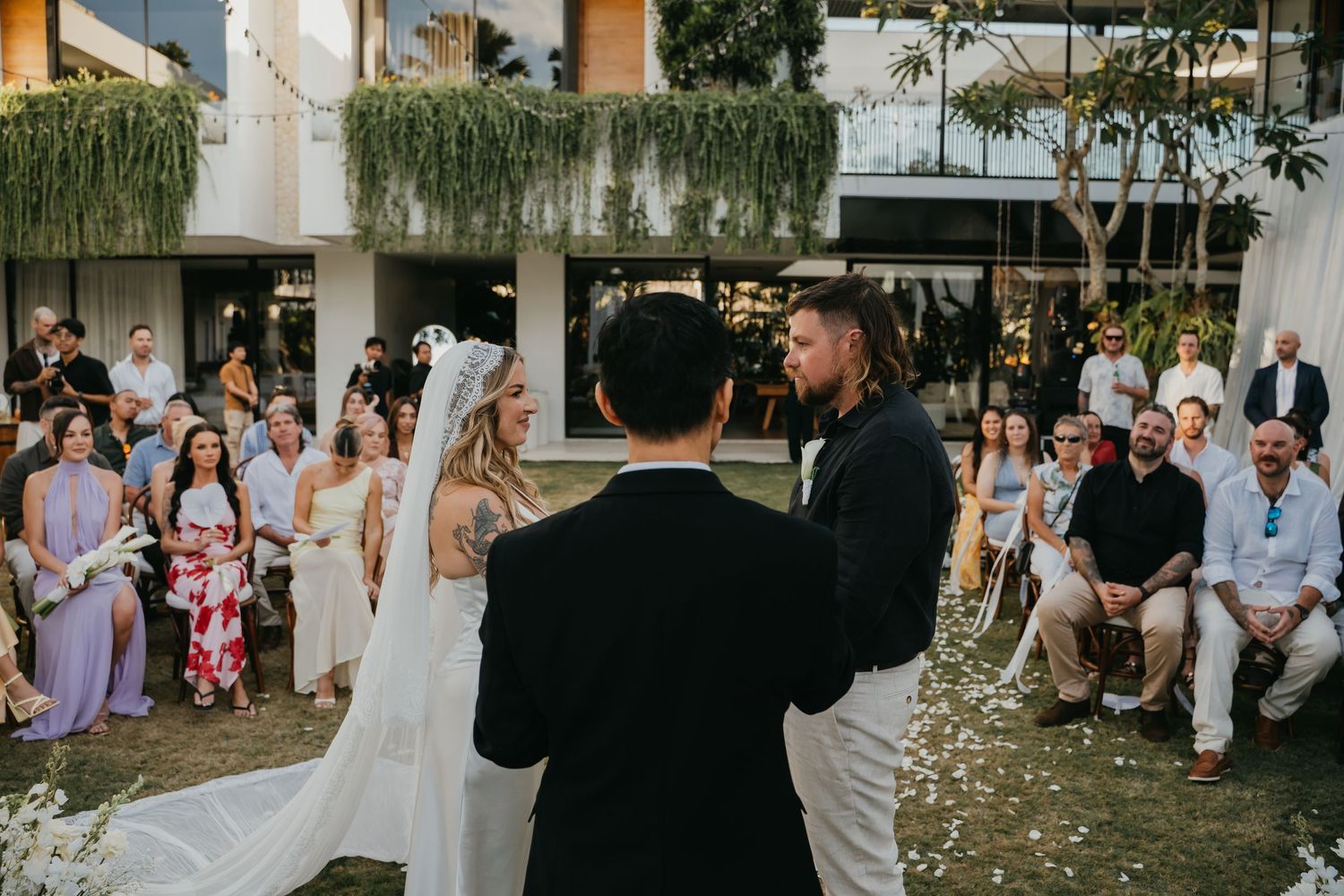 Bride and groom exchange vows before officiant at outdoor wedding ceremony with seated guests watching.