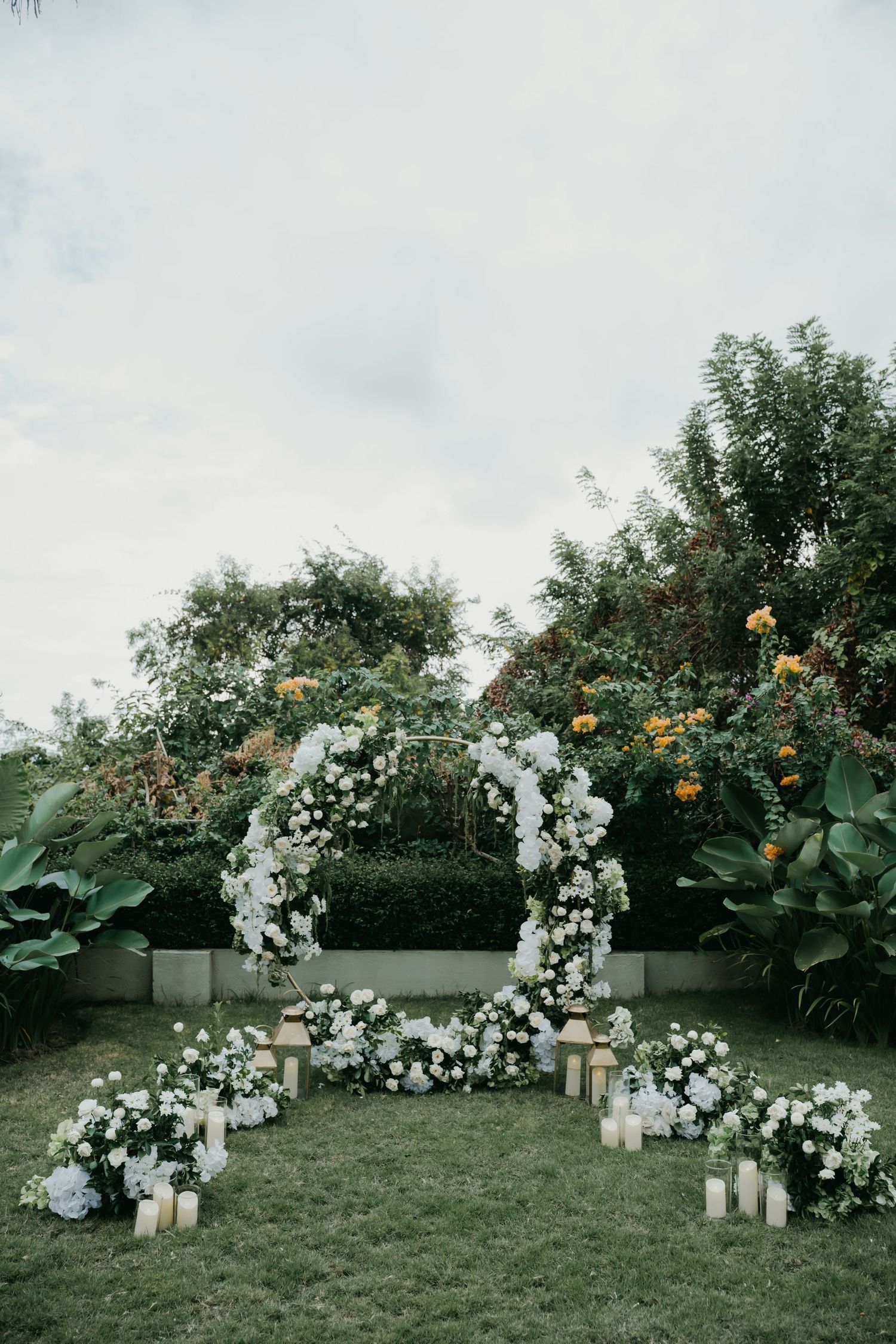 A circular white floral wedding arch with candles on green lawn surrounded by tropical gardens.