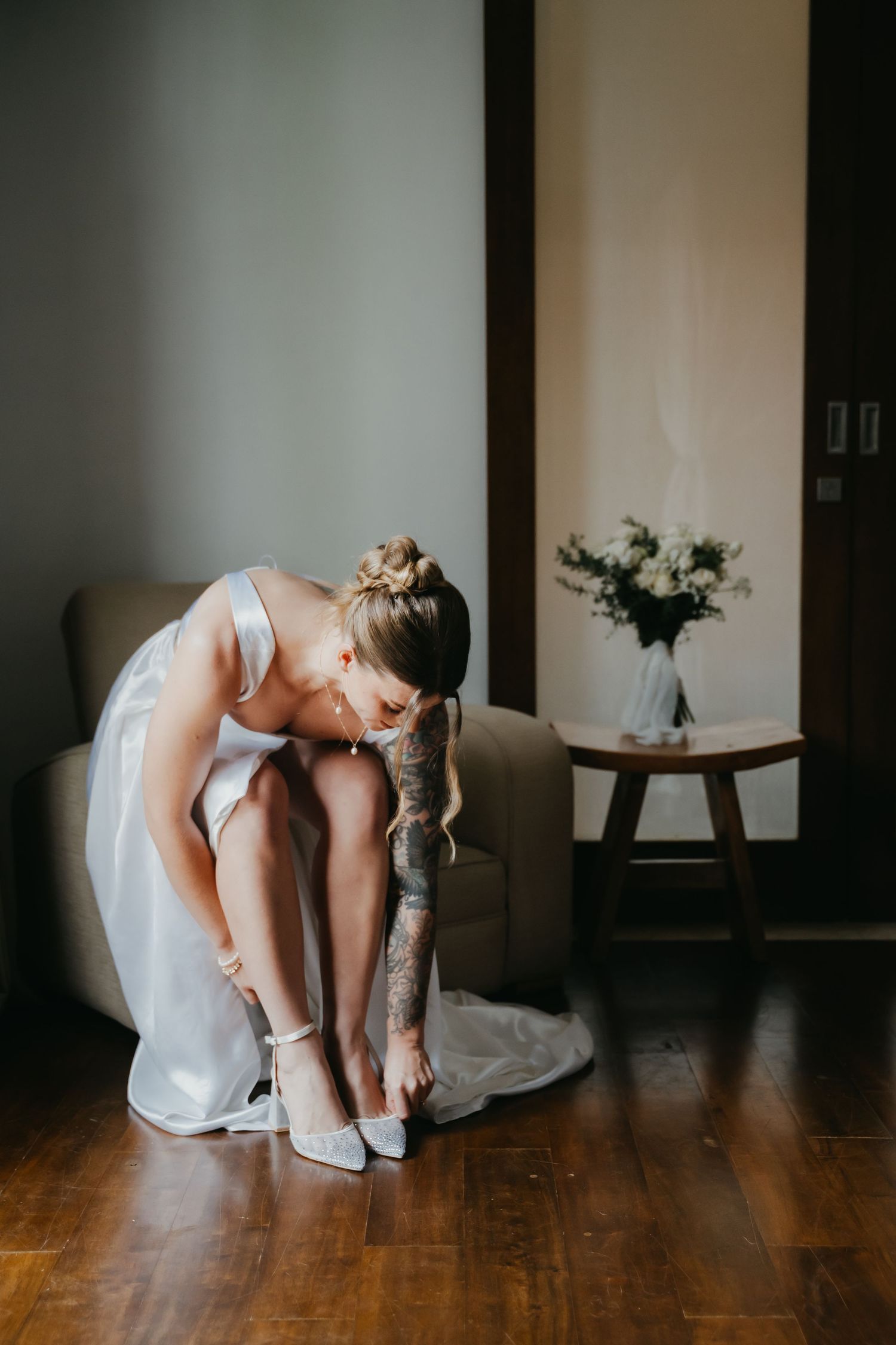Bride with tattooed arm in white wedding dress putting on heels while seated in chair near floral arrangement.