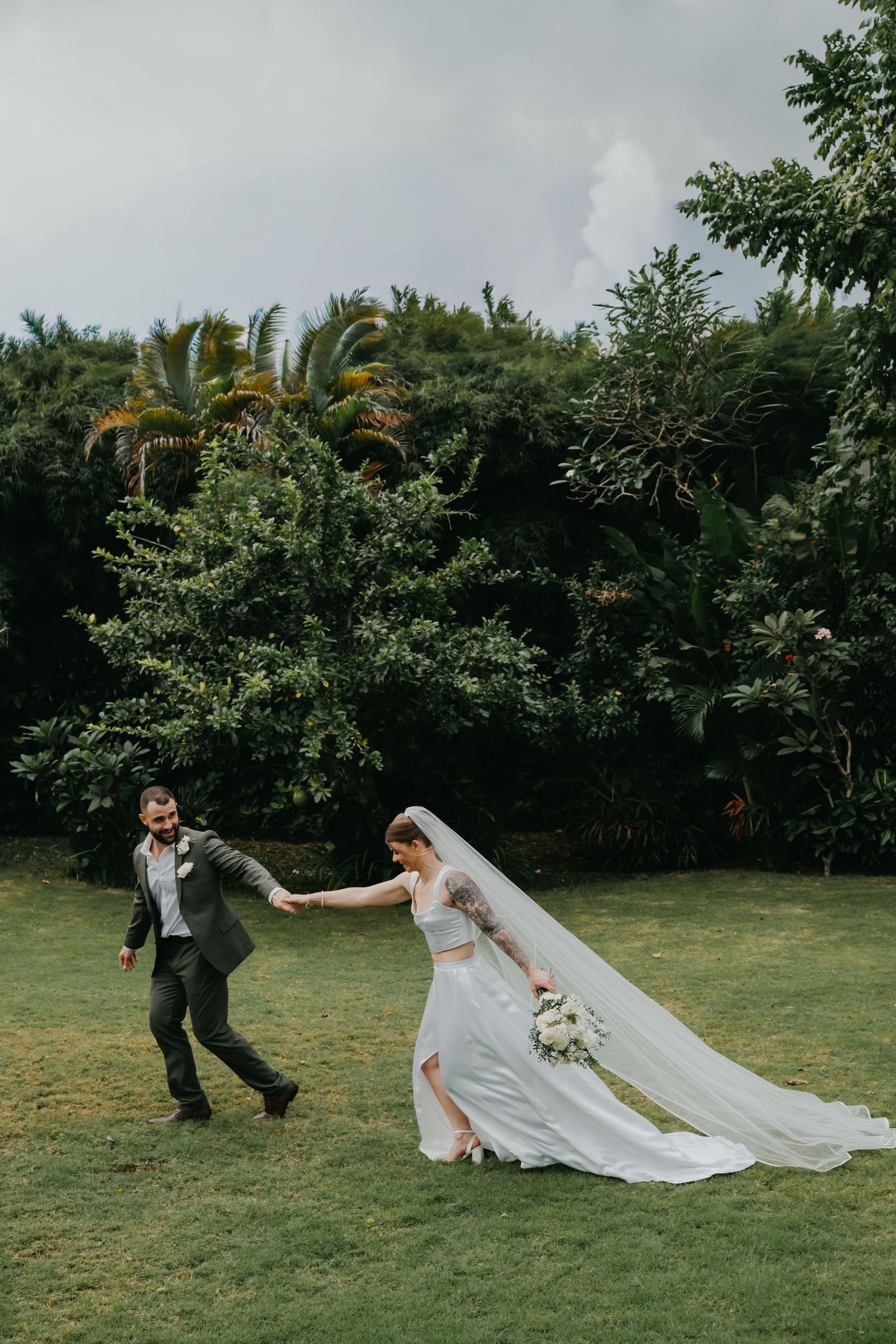Groom playfully leads bride across green lawn with her long veil flowing behind lush tropical trees.