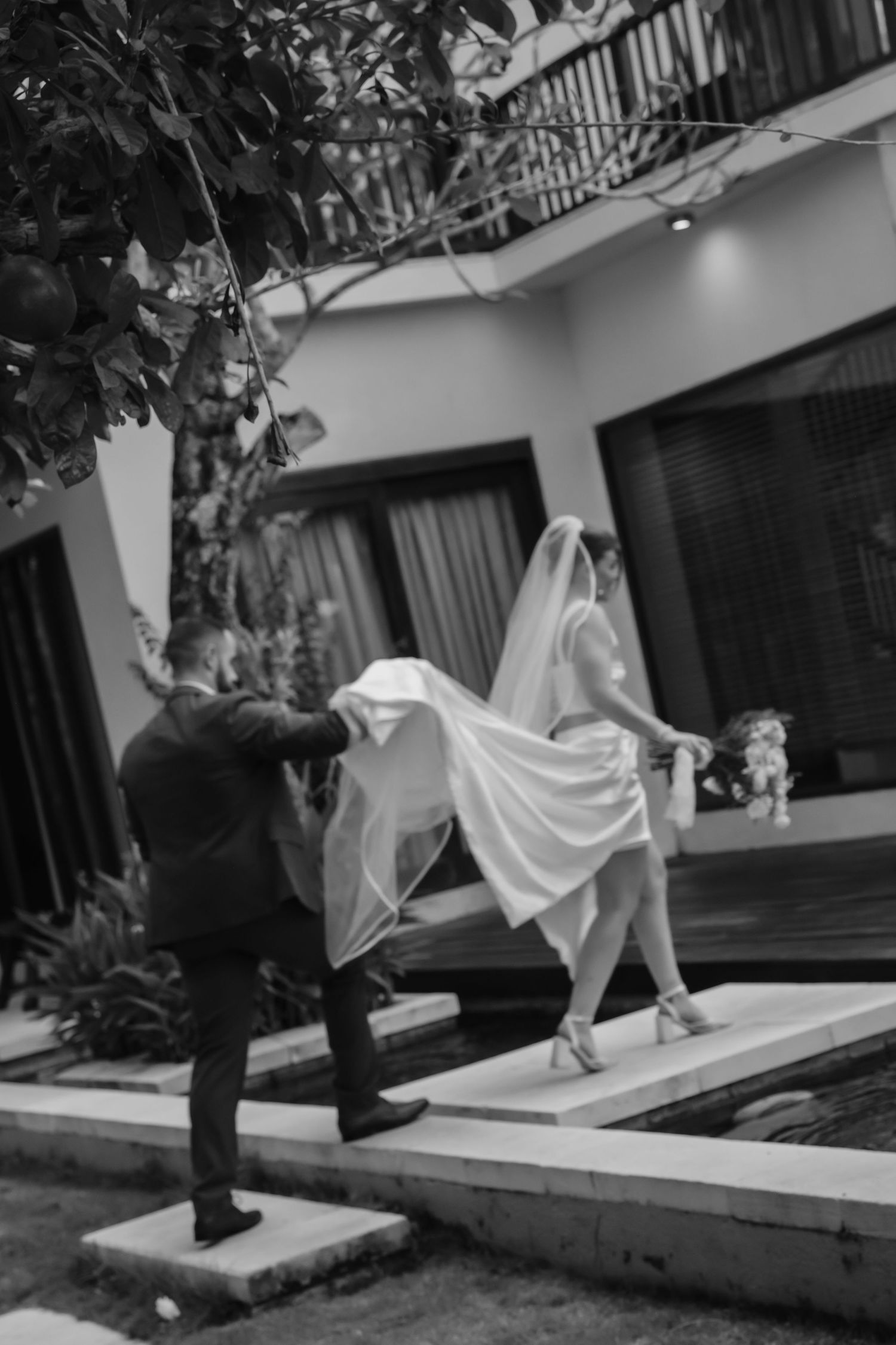 Black and white photo of groom lifting brides veil as she walks across stepping stones beside a pool.
