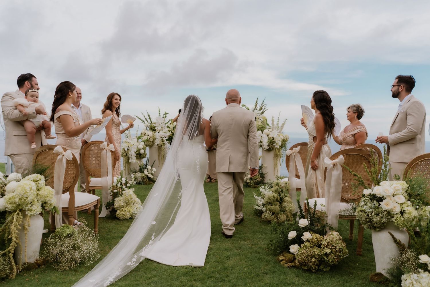 Wedding ceremony on a grassy cliff with guests, floral arrangements, and bride walking the aisle.
