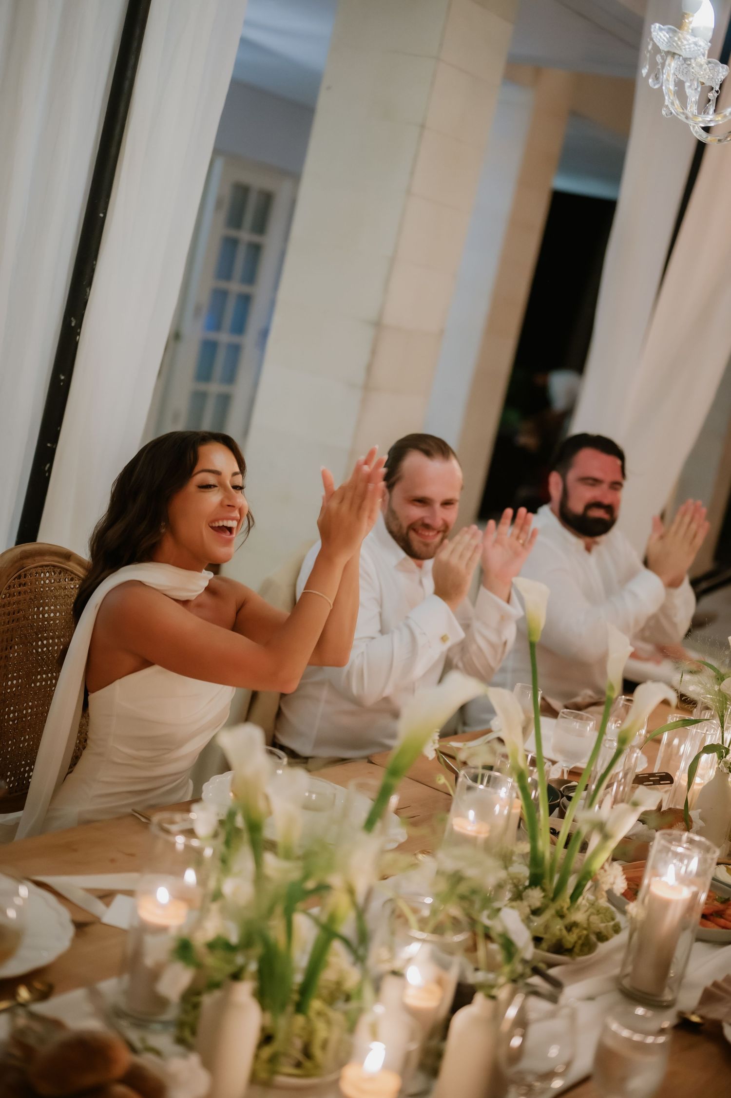 Bride and wedding guests laughing and clapping at candlelit reception dinner table with white floral centerpieces.