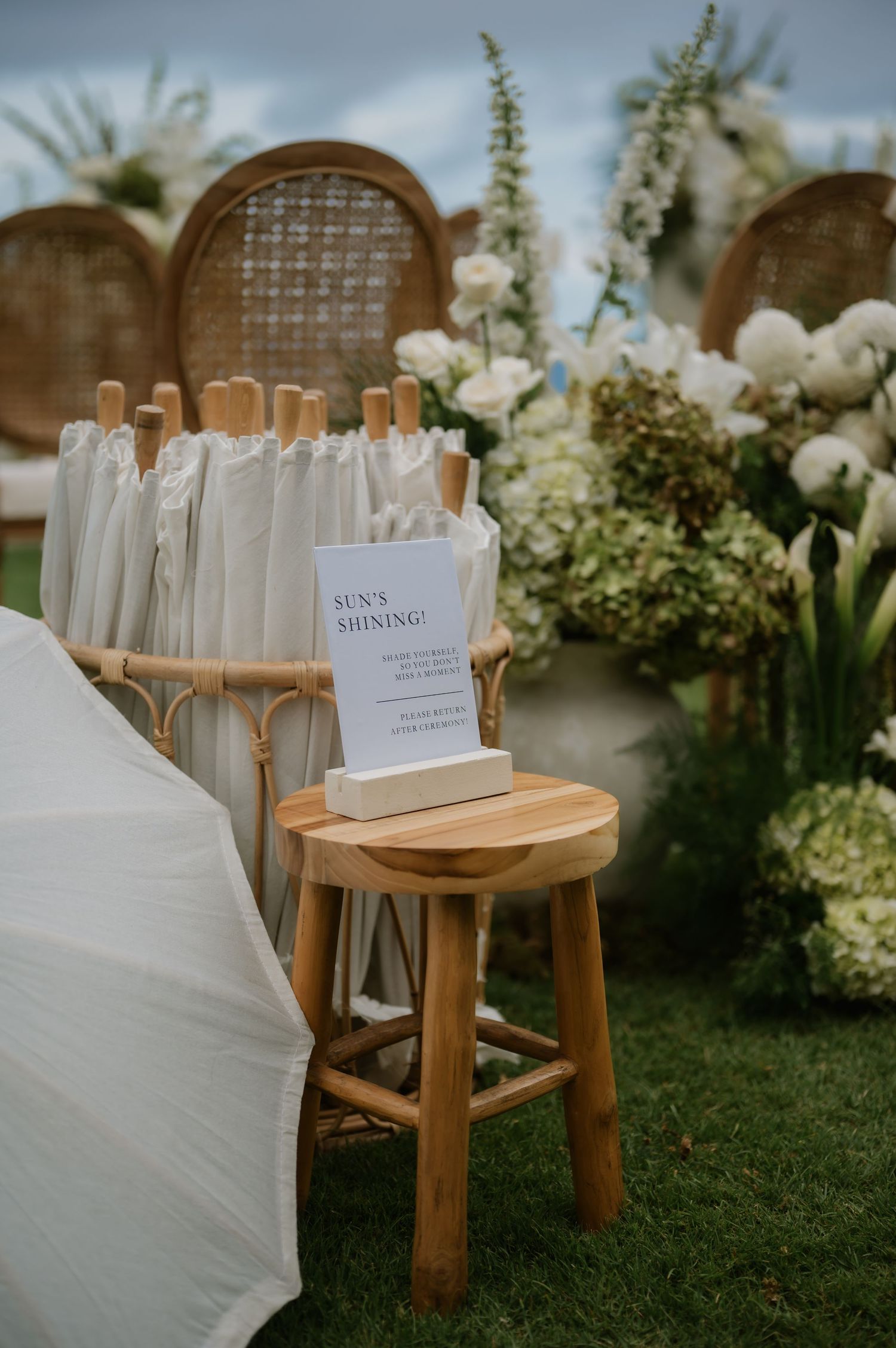 White parasols in a rattan stand beside a wooden stool with a Sun's Shining sign at an outdoor wedding ceremony.