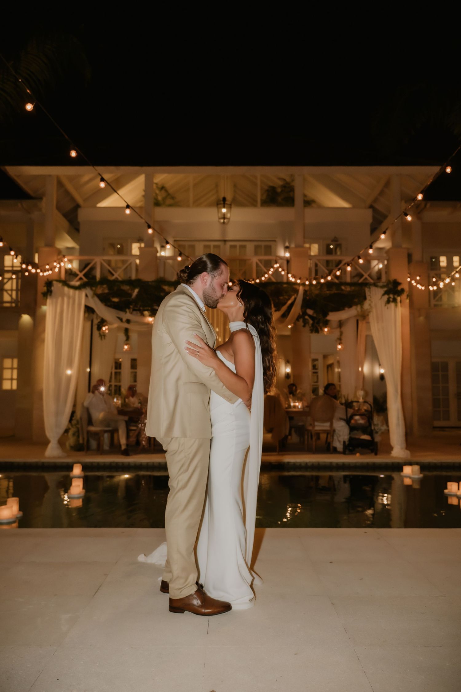 Newlyweds sharing first kiss by illuminated pool at outdoor wedding reception with string lights.