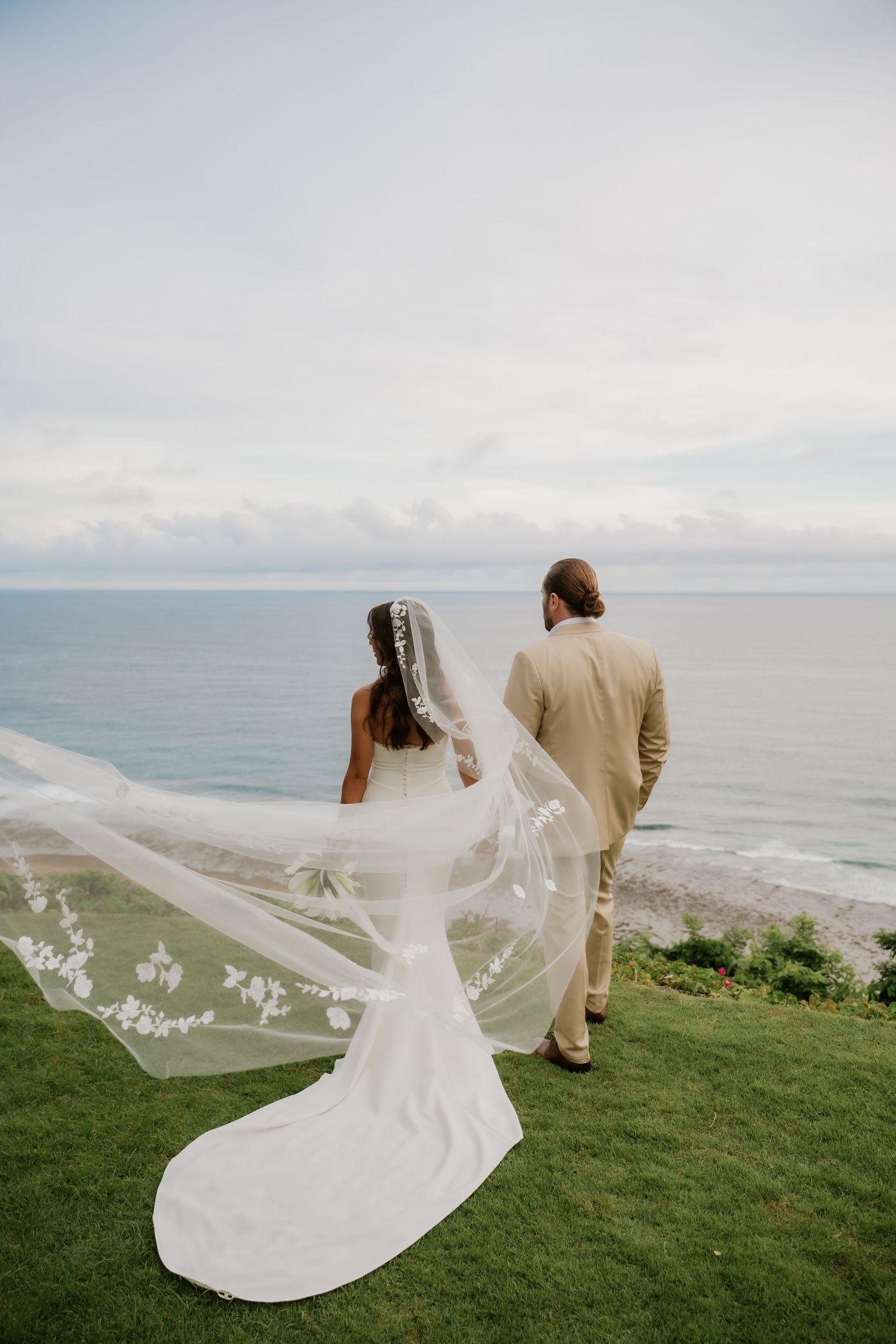 Bride and groom stand overlooking the ocean, her lace veil flowing dramatically in the breeze.