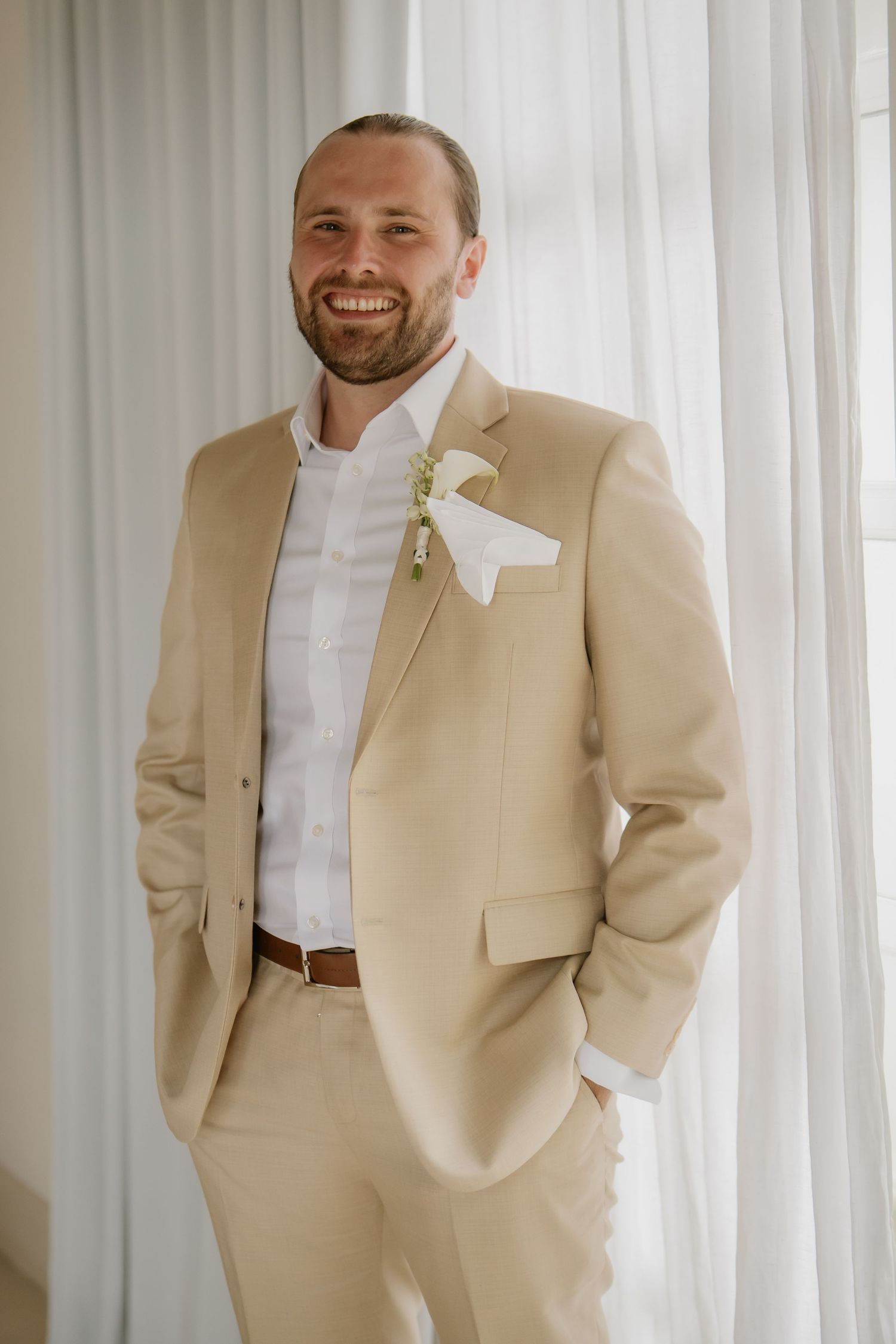 Smiling groom in a beige suit with white boutonniere and pocket square standing by sheer curtains.