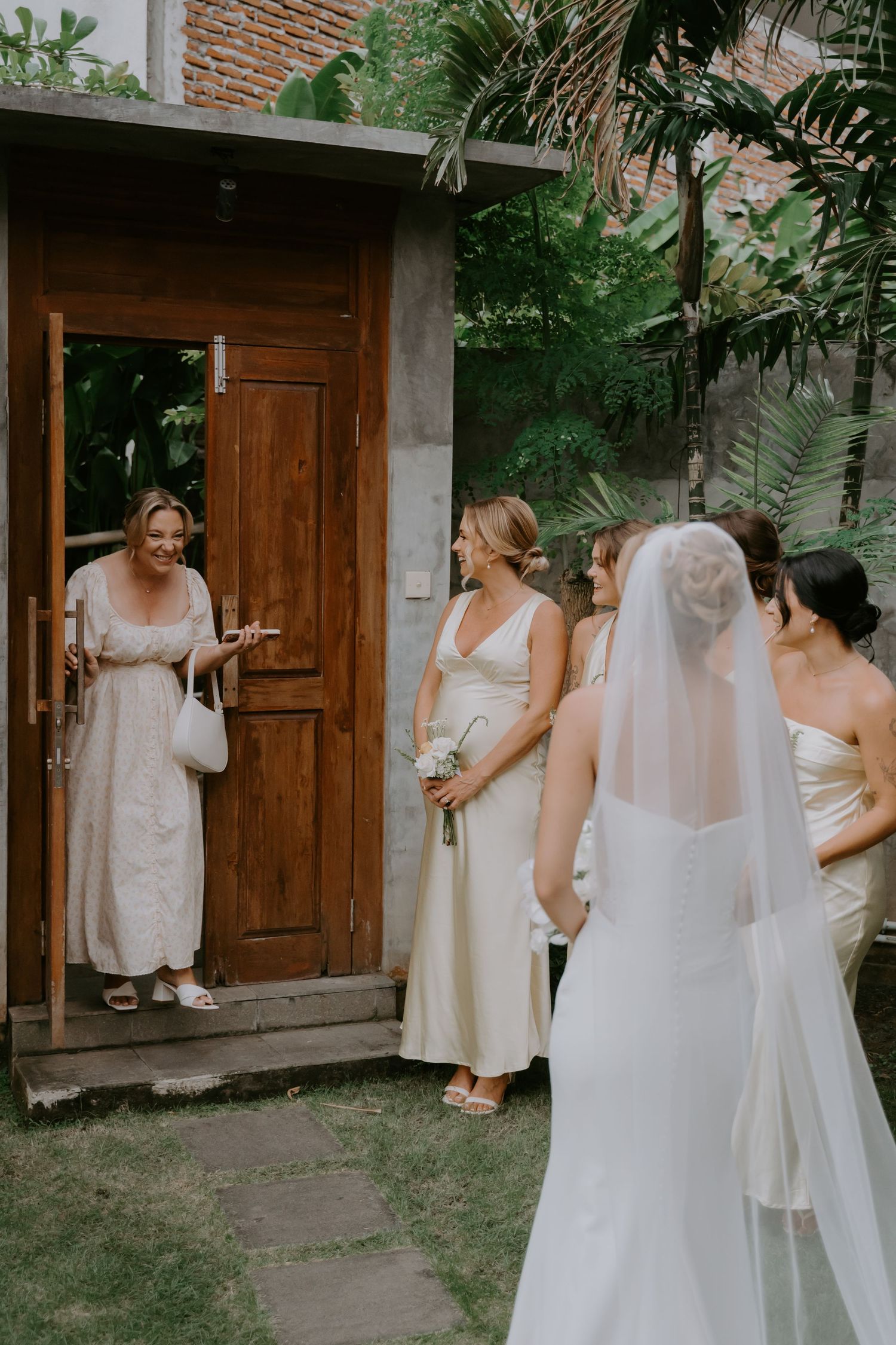 Bride in fitted white gown with veil greets guests at wooden door in garden setting.