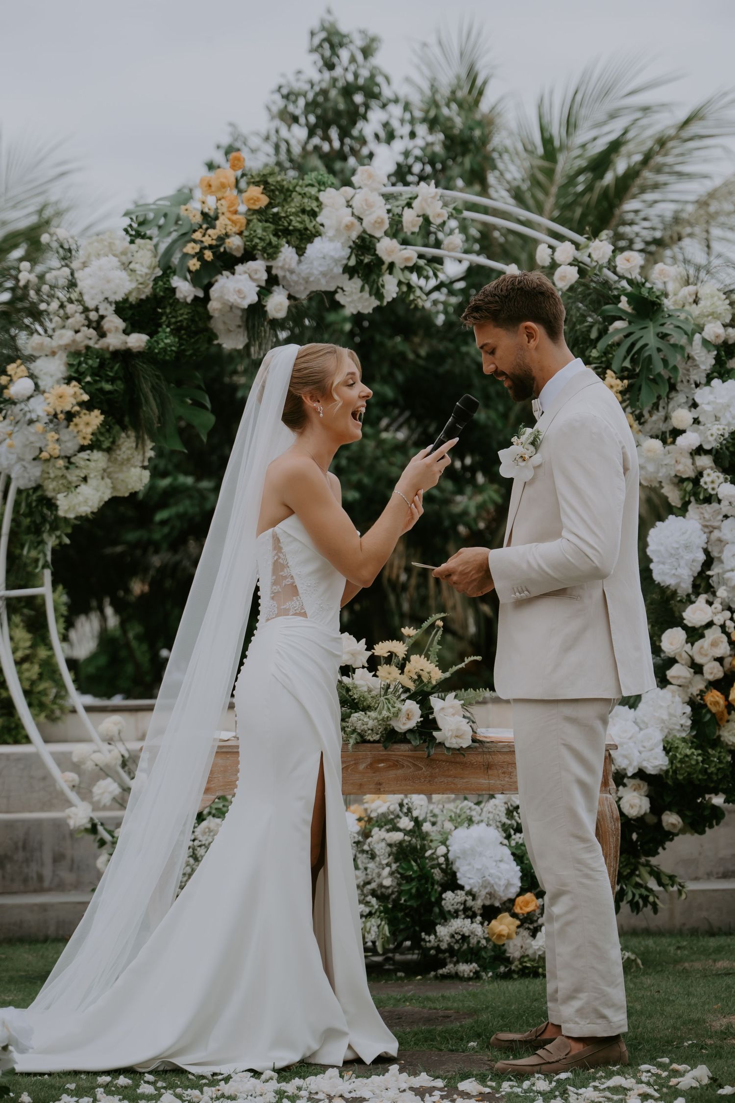 Laughing bride and groom share joyful moment during vows under lush floral wedding arch.