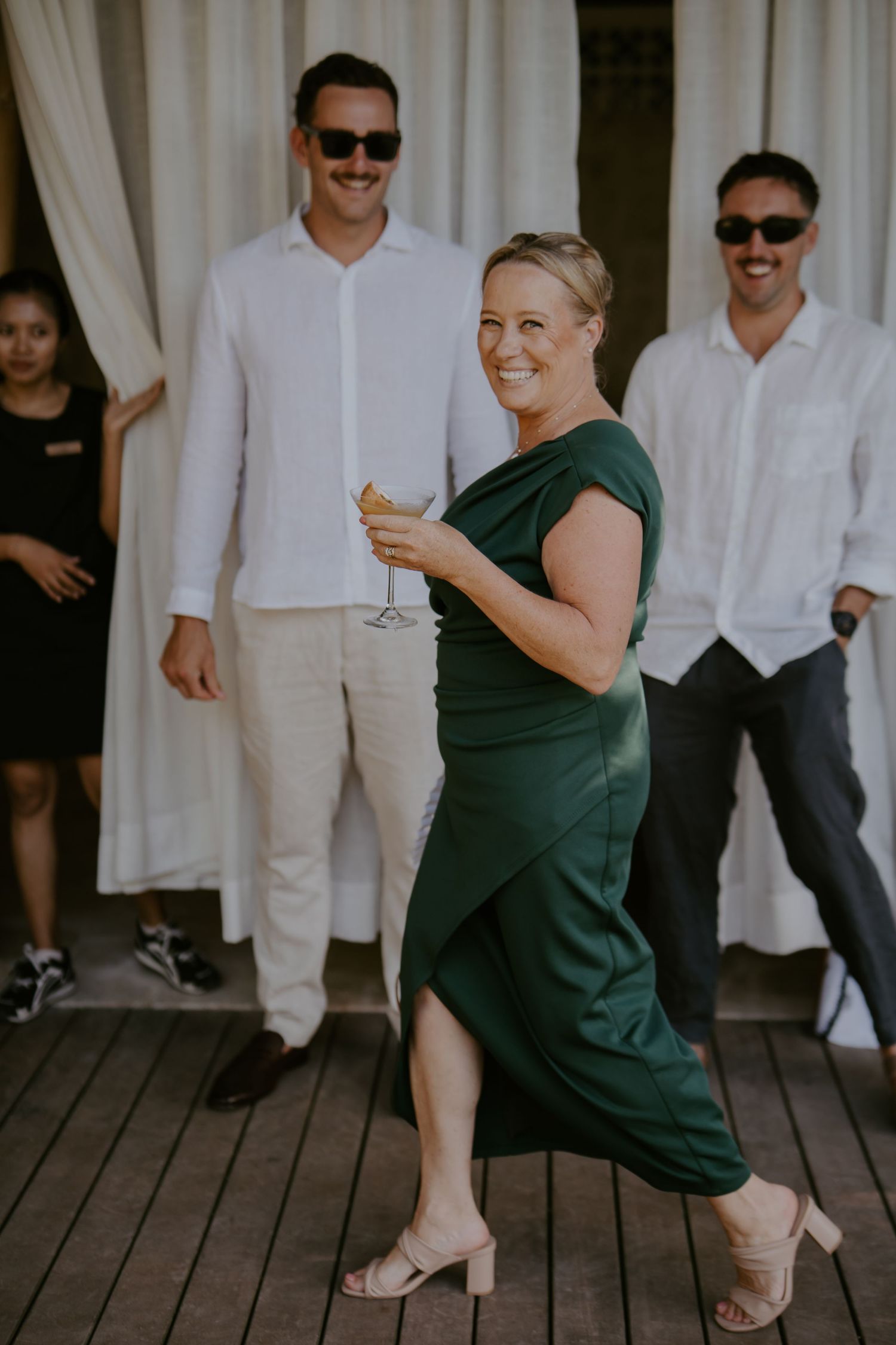 Woman in green dress holding champagne glass smiles at outdoor wedding reception on wooden deck.