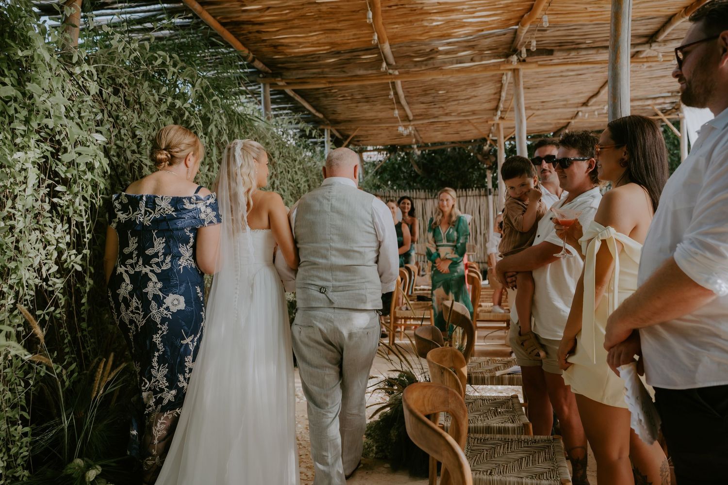 Bride in white gown walks with guests through a rustic outdoor wedding venue with greenery.