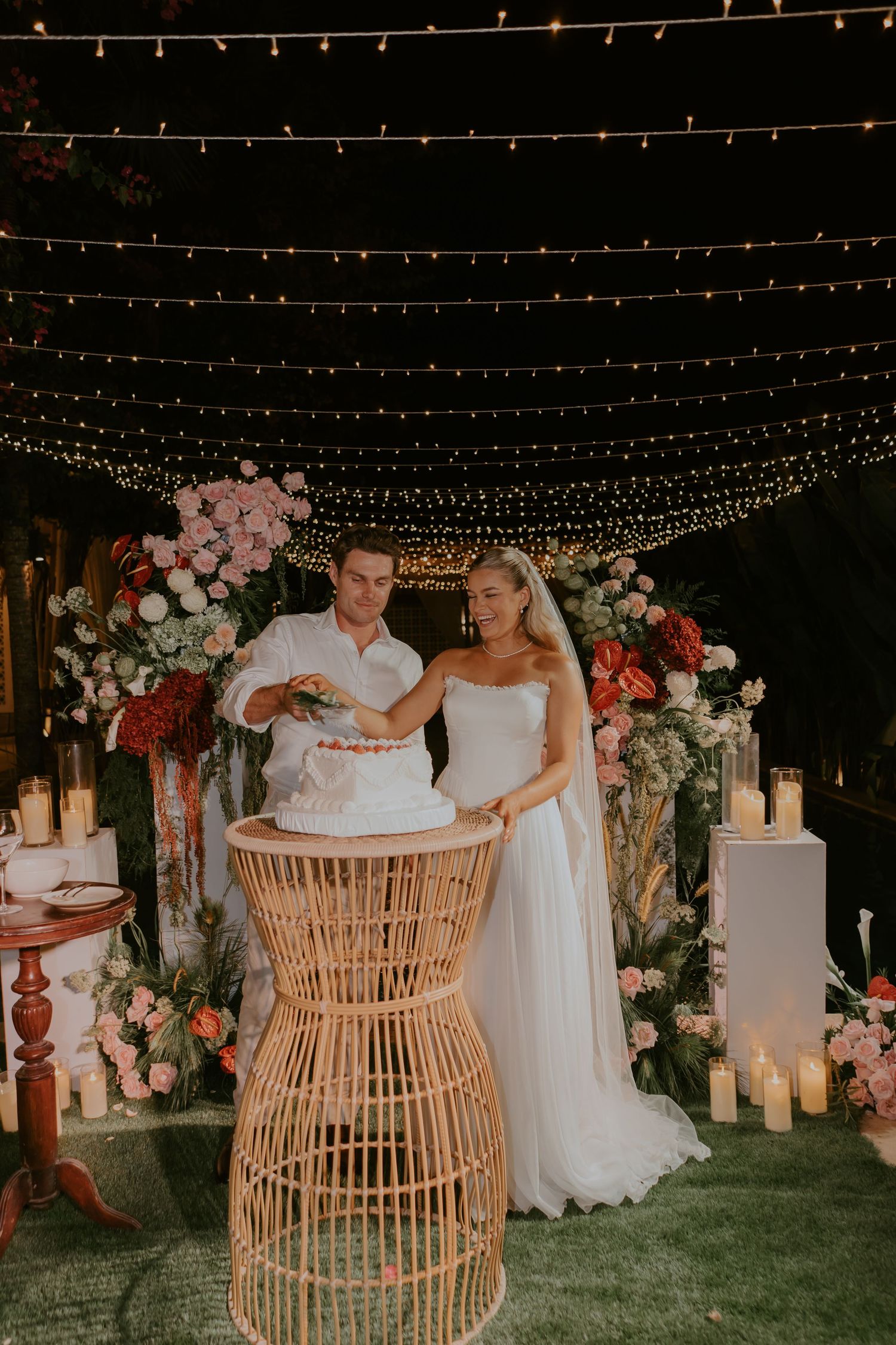 Bride and groom cutting white wedding cake surrounded by candles and floral arrangements under string lights.