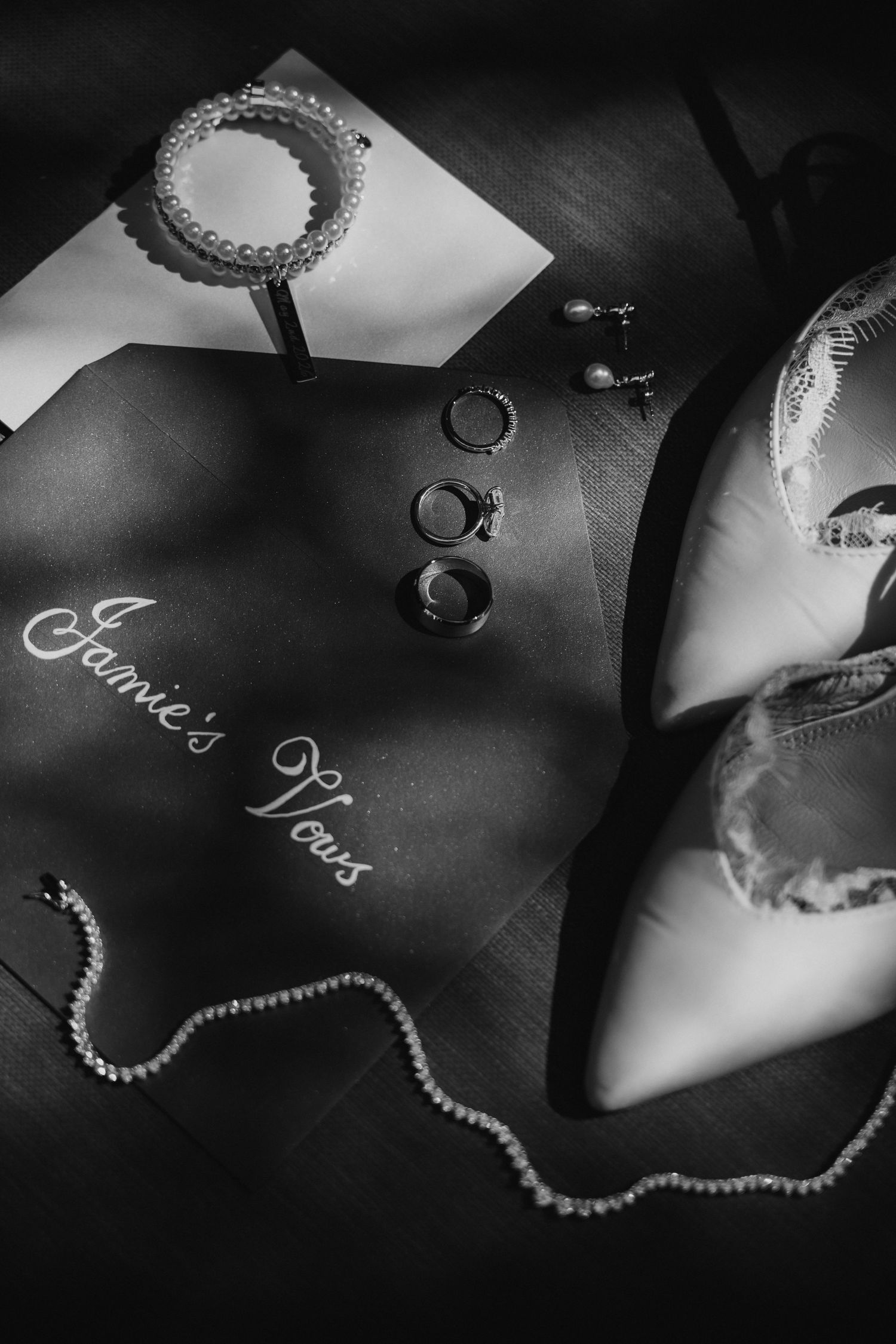 Black and white flat lay of Jamie's wedding vows book, rings, pearl necklace, earrings, and bridal heels.