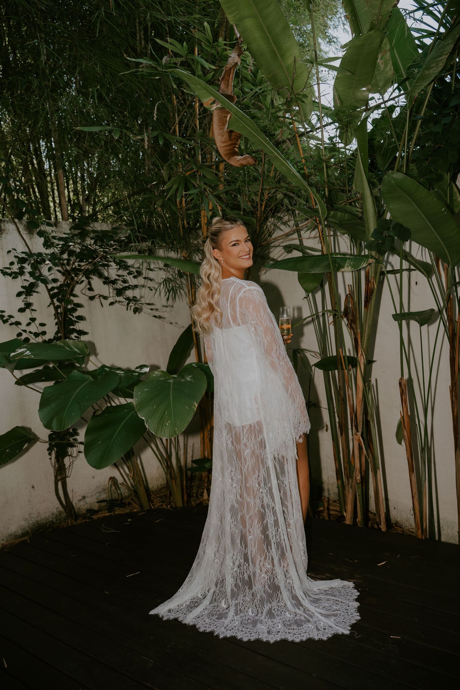 Bride in elegant white lace cape gown posing among tropical greenery on her wedding day.