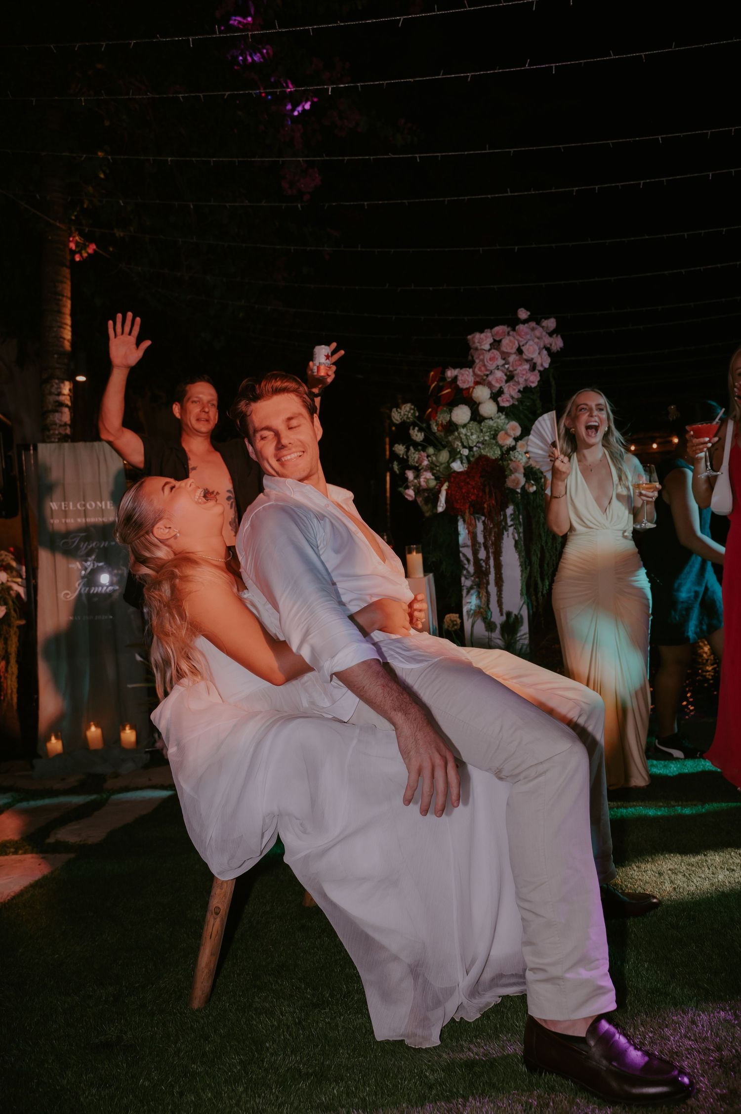 Laughing bride sits on grooms lap during fun wedding reception moment with cheering guests behind them.
