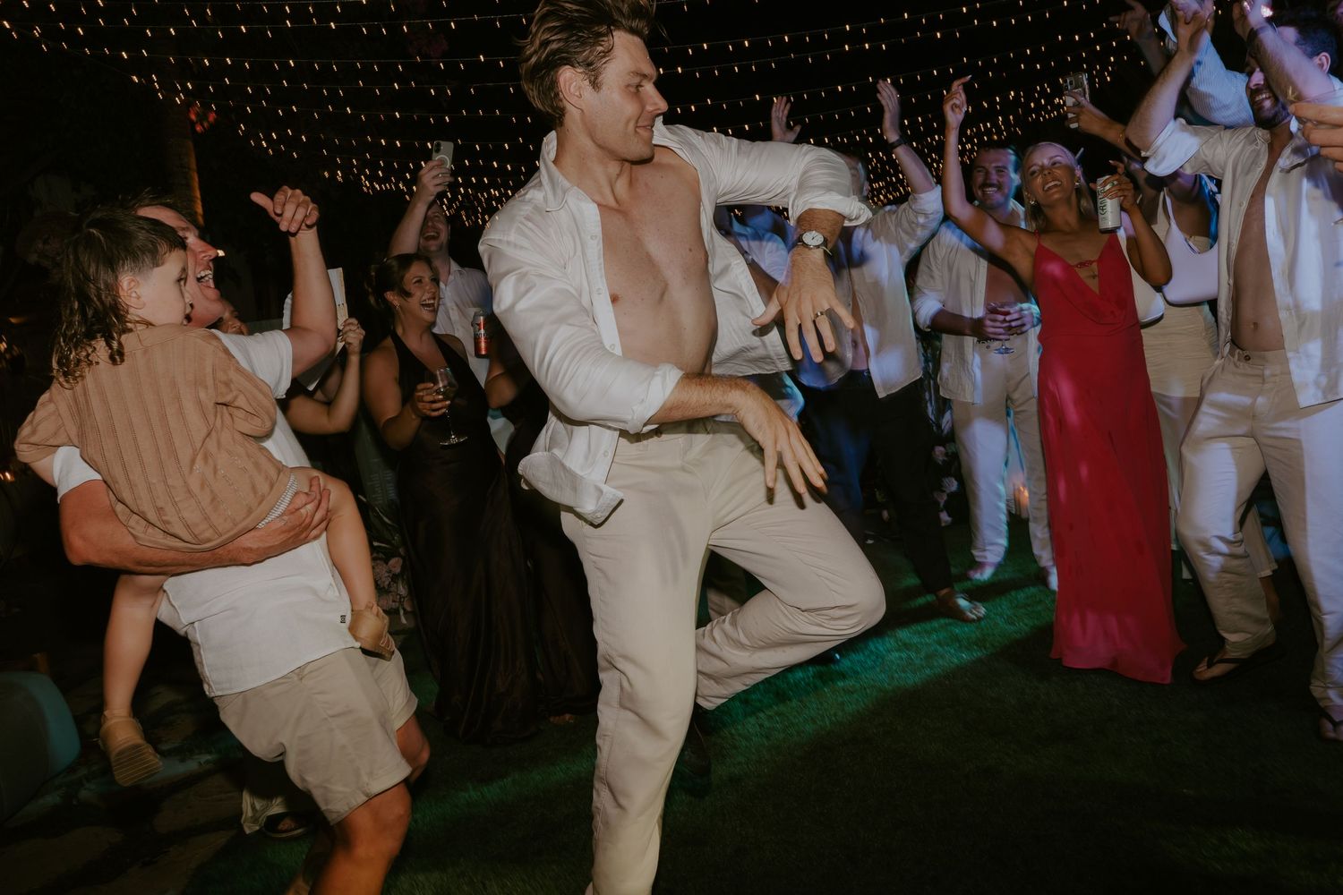 Shirtless groom dancing energetically with guests under string lights at lively wedding reception.