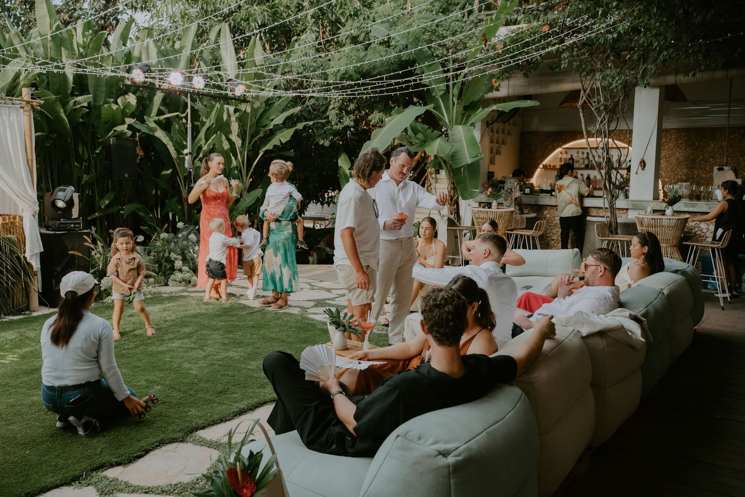 Wedding reception guests mingle and relax in a lush tropical outdoor garden venue decorated with string lights.