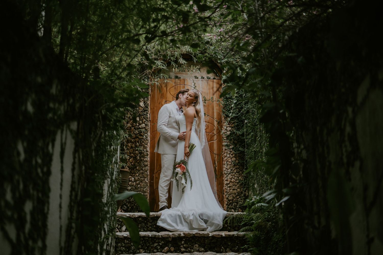 Bride and groom share romantic kiss in lush green garden pathway at outdoor wedding ceremony.