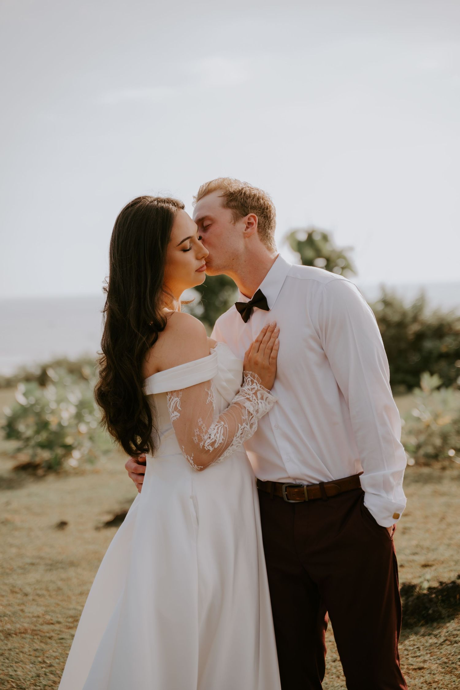 Bride and groom share a romantic kiss outdoors at sunset with ocean views behind them.