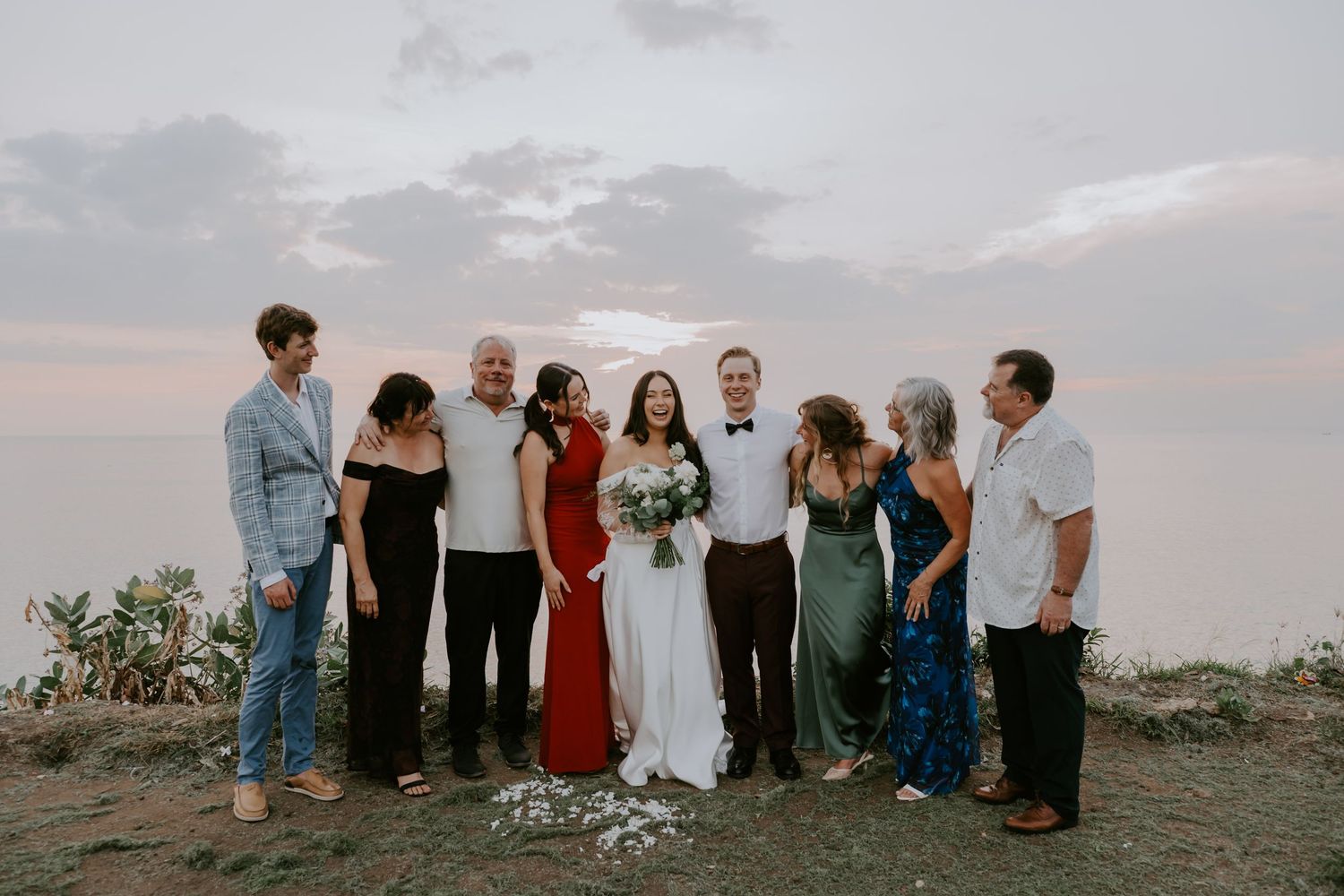 Wedding party poses together overlooking dramatic ocean sunset on clifftop venue.
