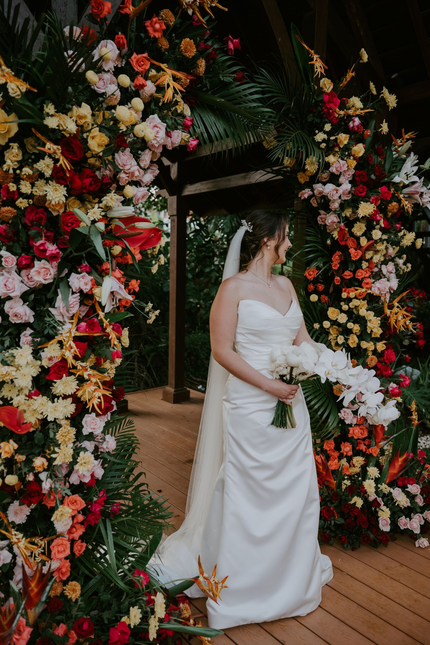 Bride in strapless white gown holding white bouquet stands beside vibrant tropical floral wedding arch.