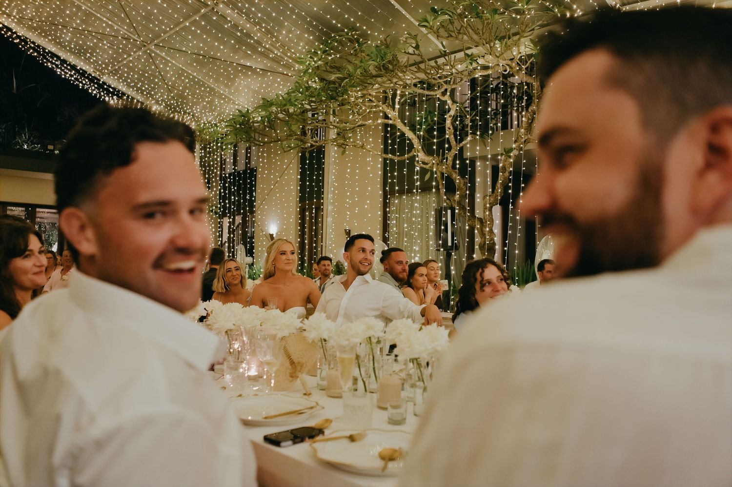 Two smiling groomsmen in white shirts sit at reception table under canopy of twinkling fairy lights.