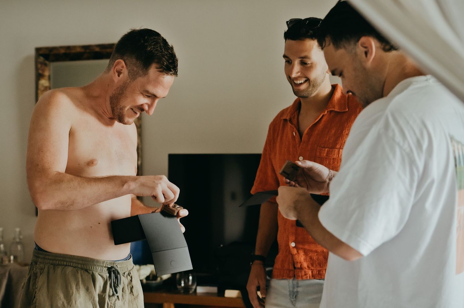 Three men laughing together indoors, one shirtless holding an item, others in casual shirts in a bright room.