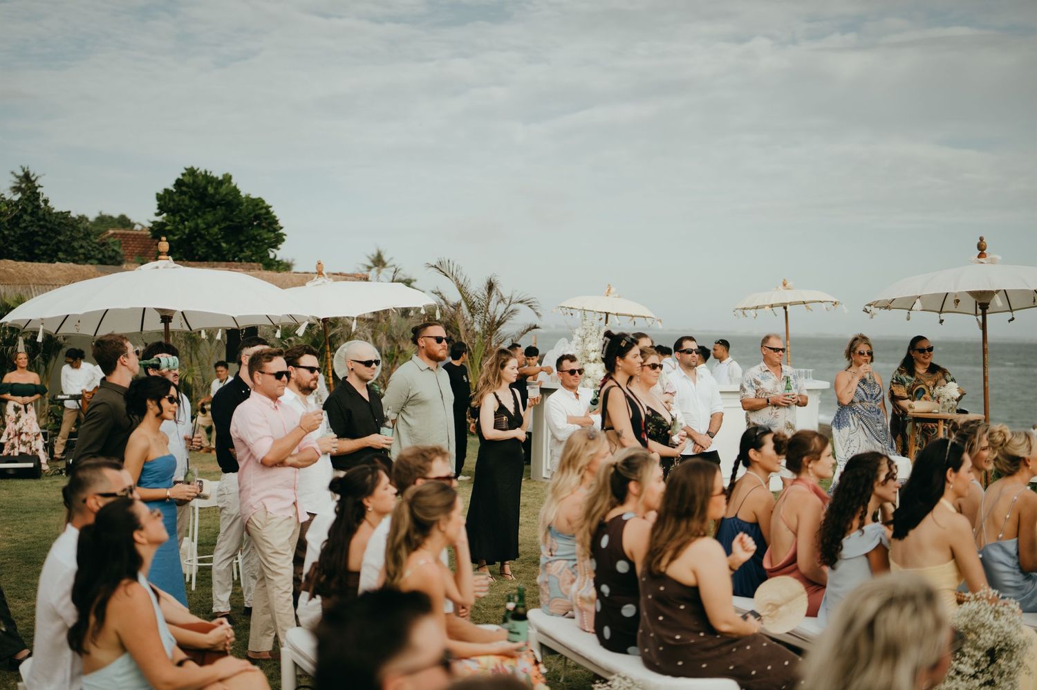 Wedding guests gathered outdoors under white umbrellas at oceanfront ceremony venue in Bali.