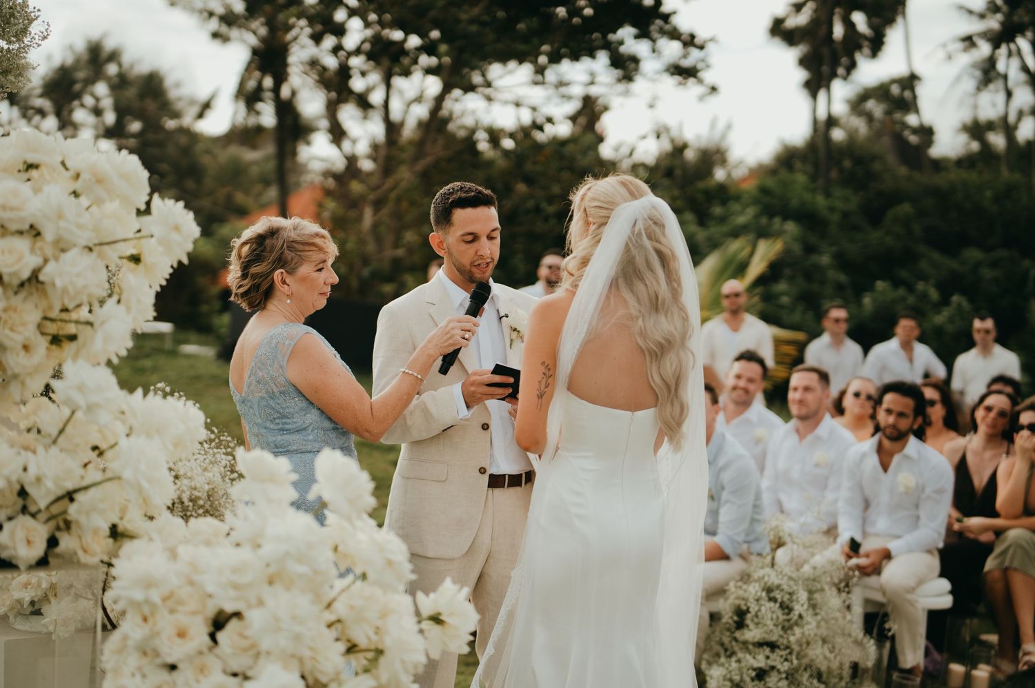 Groom in beige suit exchanges vows with bride at floral-adorned outdoor wedding ceremony.