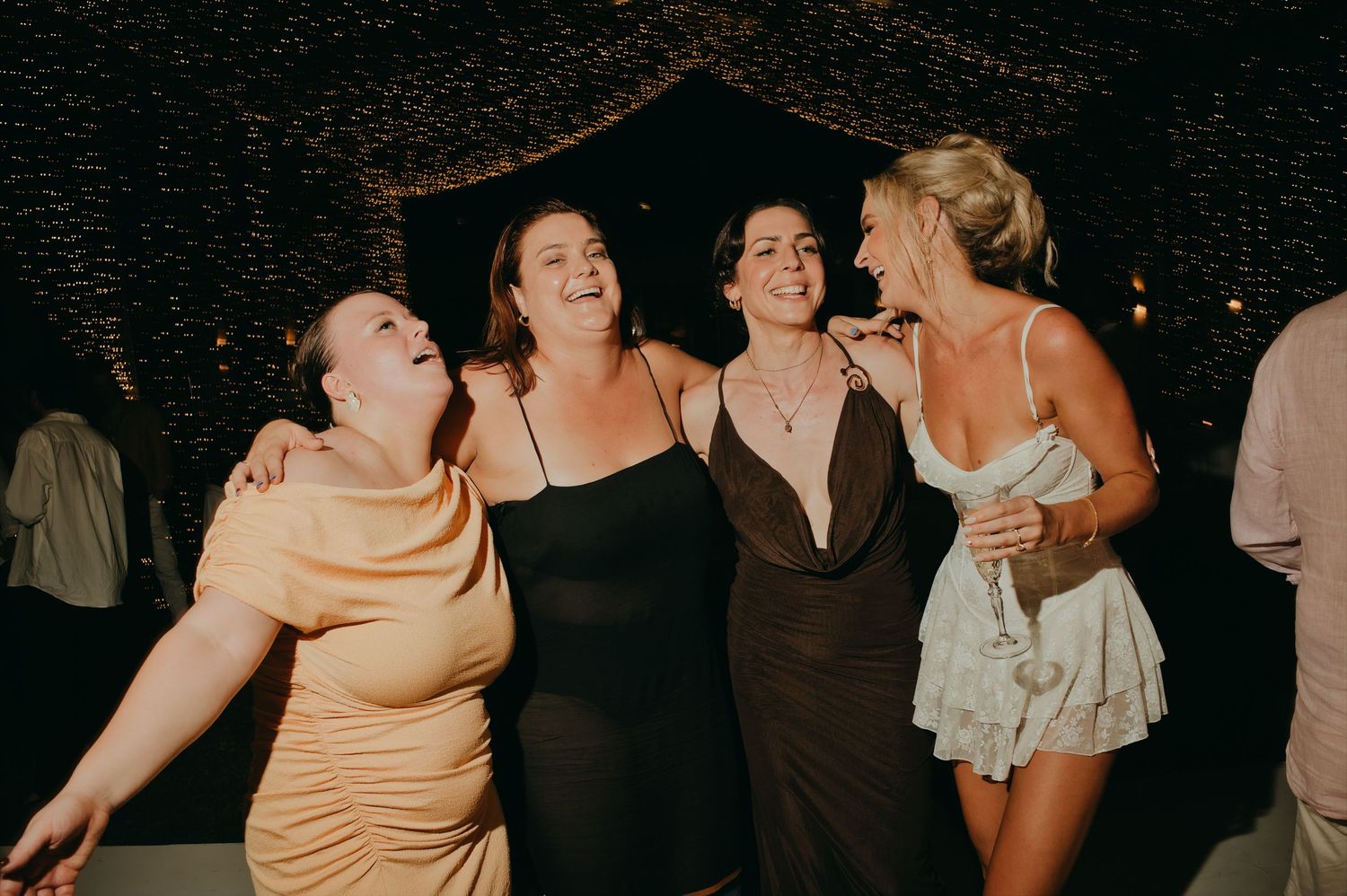 Four women laugh and embrace on the dance floor at a festive wedding reception at night.