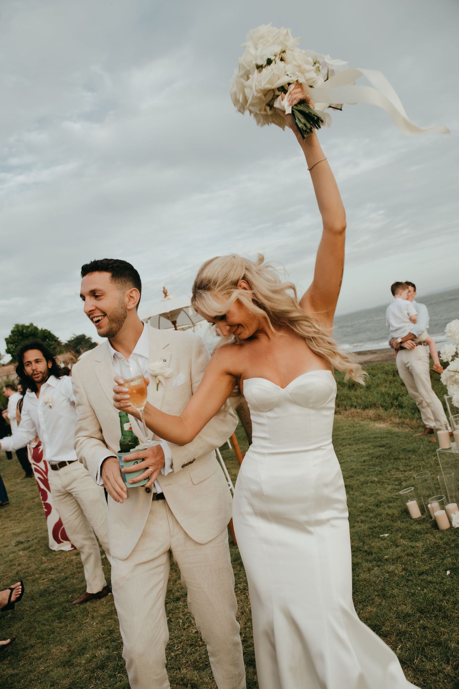 Joyful bride in white gown raises bouquet while groom in beige suit celebrates after outdoor wedding ceremony.