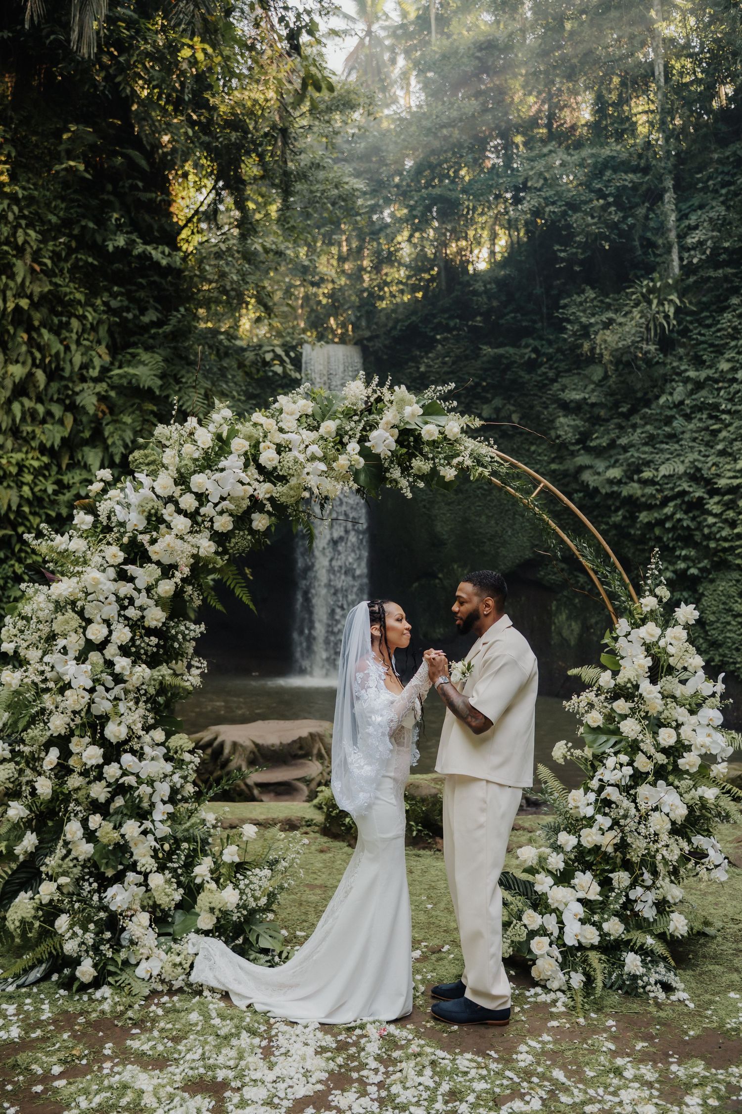 Bride and groom exchange vows under a floral arch with a waterfall backdrop in a lush tropical forest.