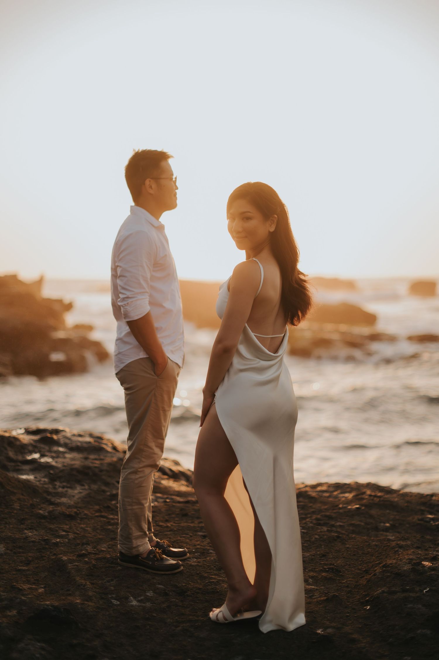 Couple posing at golden hour on rocky shore, woman in white backless gown holding hands with man in white shirt.