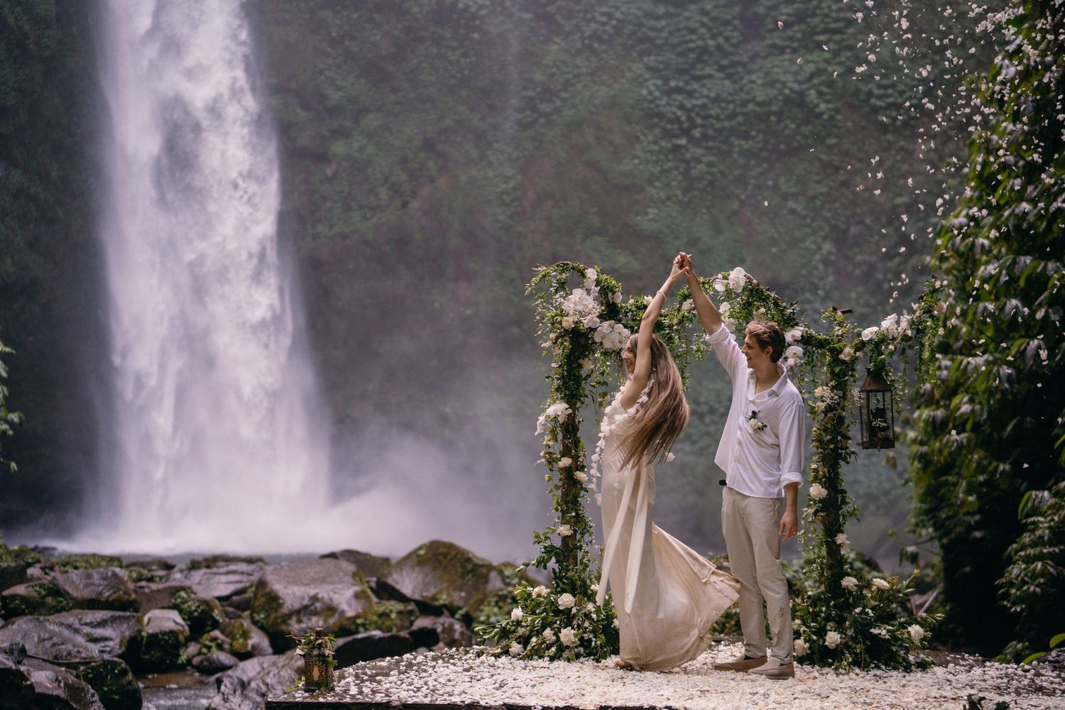 A couple celebrates their wedding ceremony under a floral arch beside a majestic waterfall in a lush natural setting.