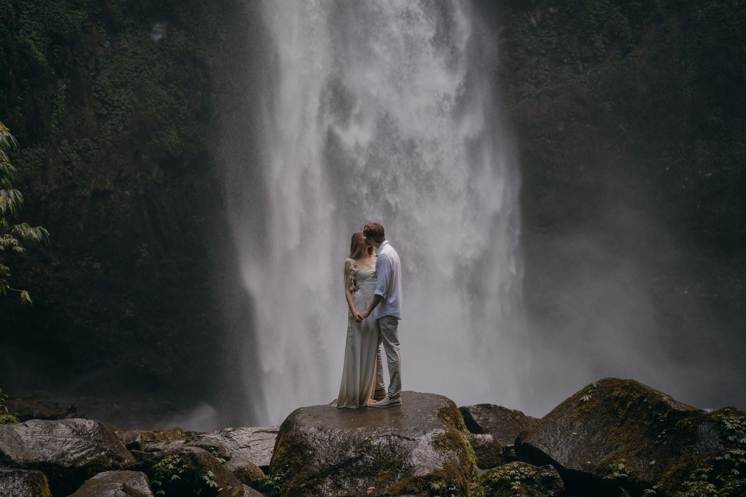 A couple embraces on a rocky outcrop in front of a majestic cascading waterfall in a lush forest setting.