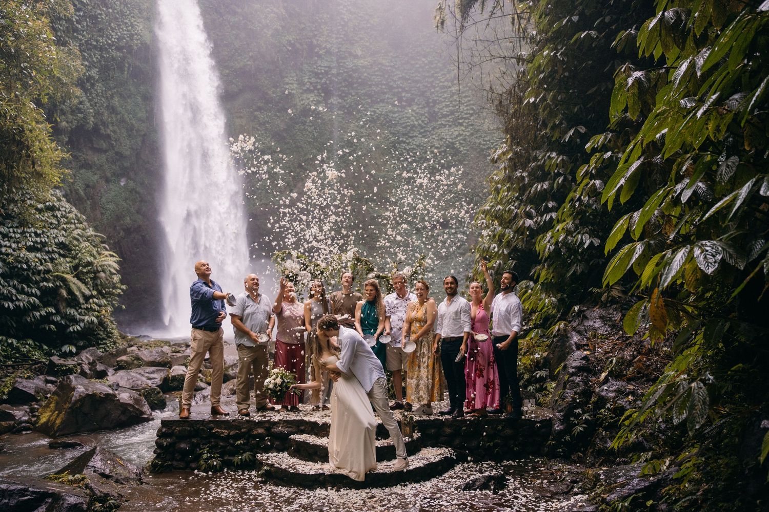 A wedding ceremony takes place in front of a majestic waterfall surrounded by lush tropical foliage.