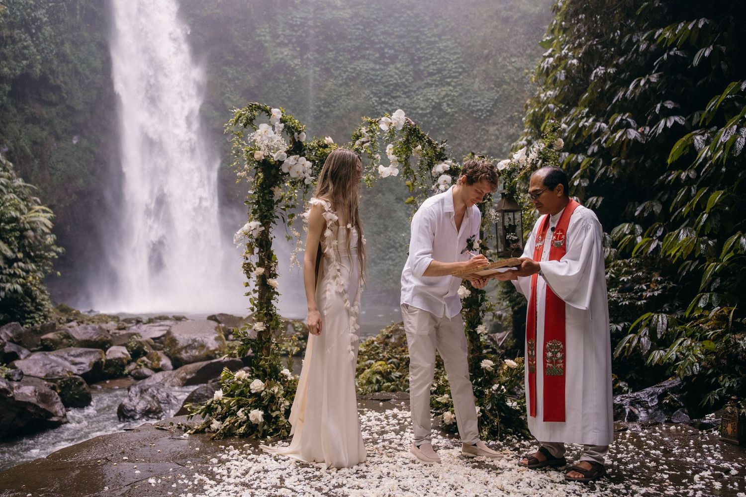 A wedding ceremony takes place in front of a majestic waterfall with floral arch decorations in a lush tropical setting.