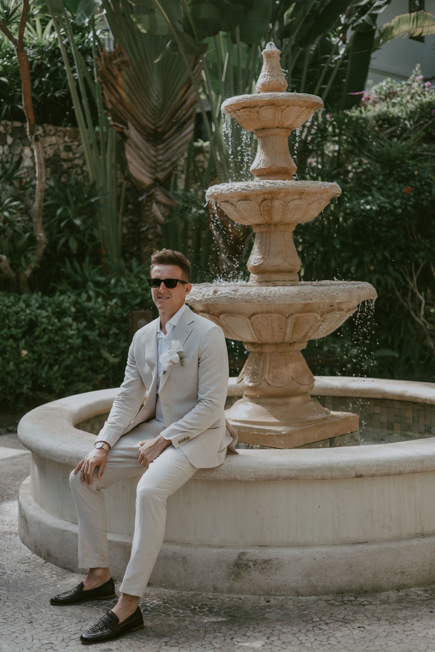 Person in light colored suit sits elegantly by ornate stone fountain in garden setting.