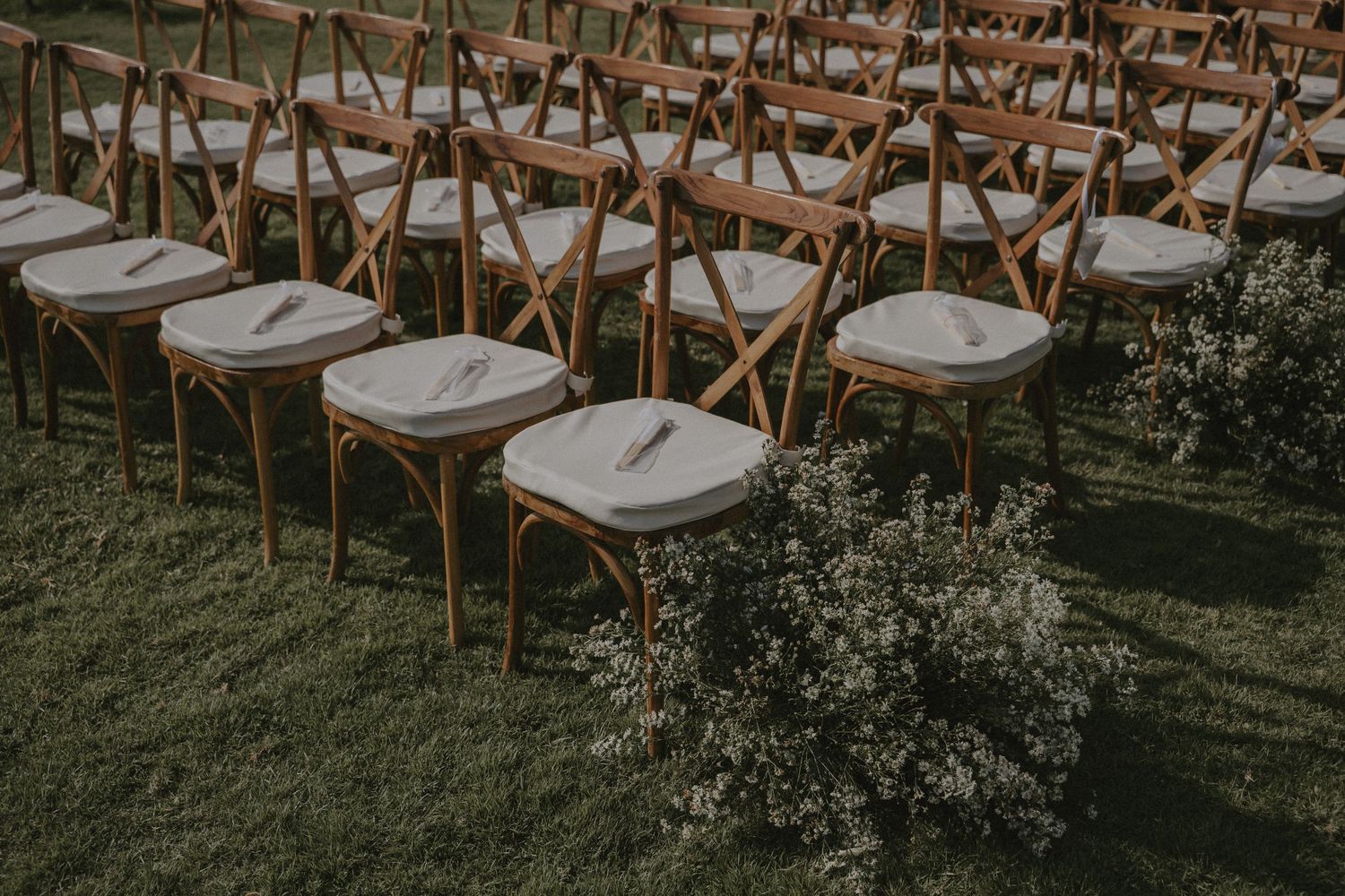 Rows of rustic wooden crossback chairs arranged on a green lawn for an elegant outdoor wedding ceremony.