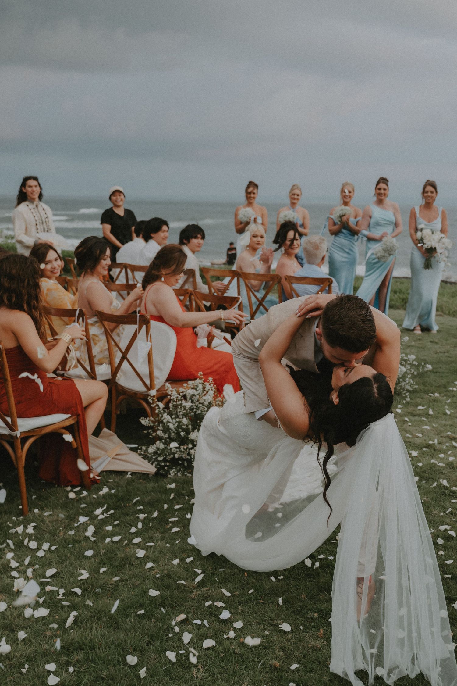 Wedding party members in white and coral dresses pose playfully on a grassy lawn overlooking the ocean.