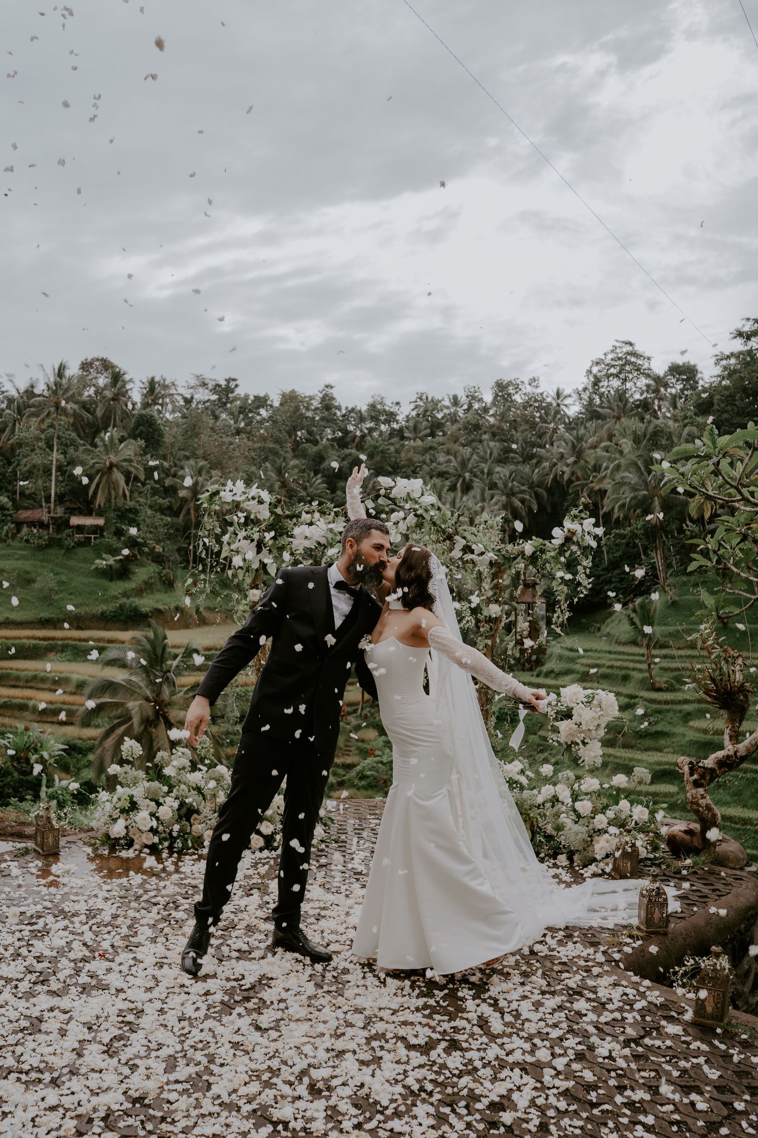 A couple celebrates their wedding day in an orchard as flower petals float through the air during a joyful outdoor celebration.