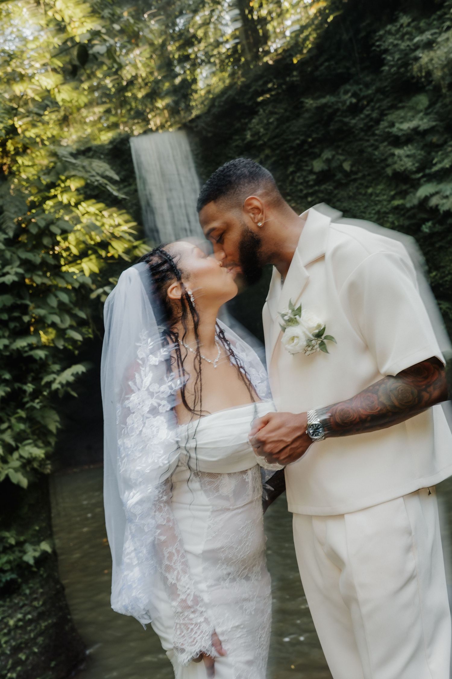Interracial couple shares a romantic kiss in a tropical garden setting with a waterfall backdrop on their wedding day.