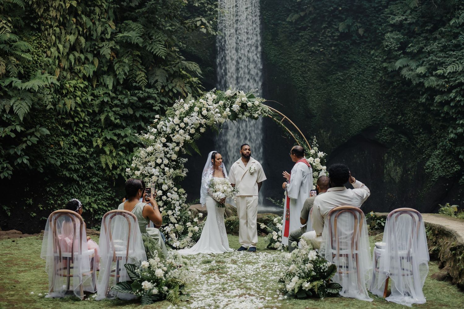 Outdoor wedding ceremony with floral arch and waterfall backdrop in lush green garden setting.